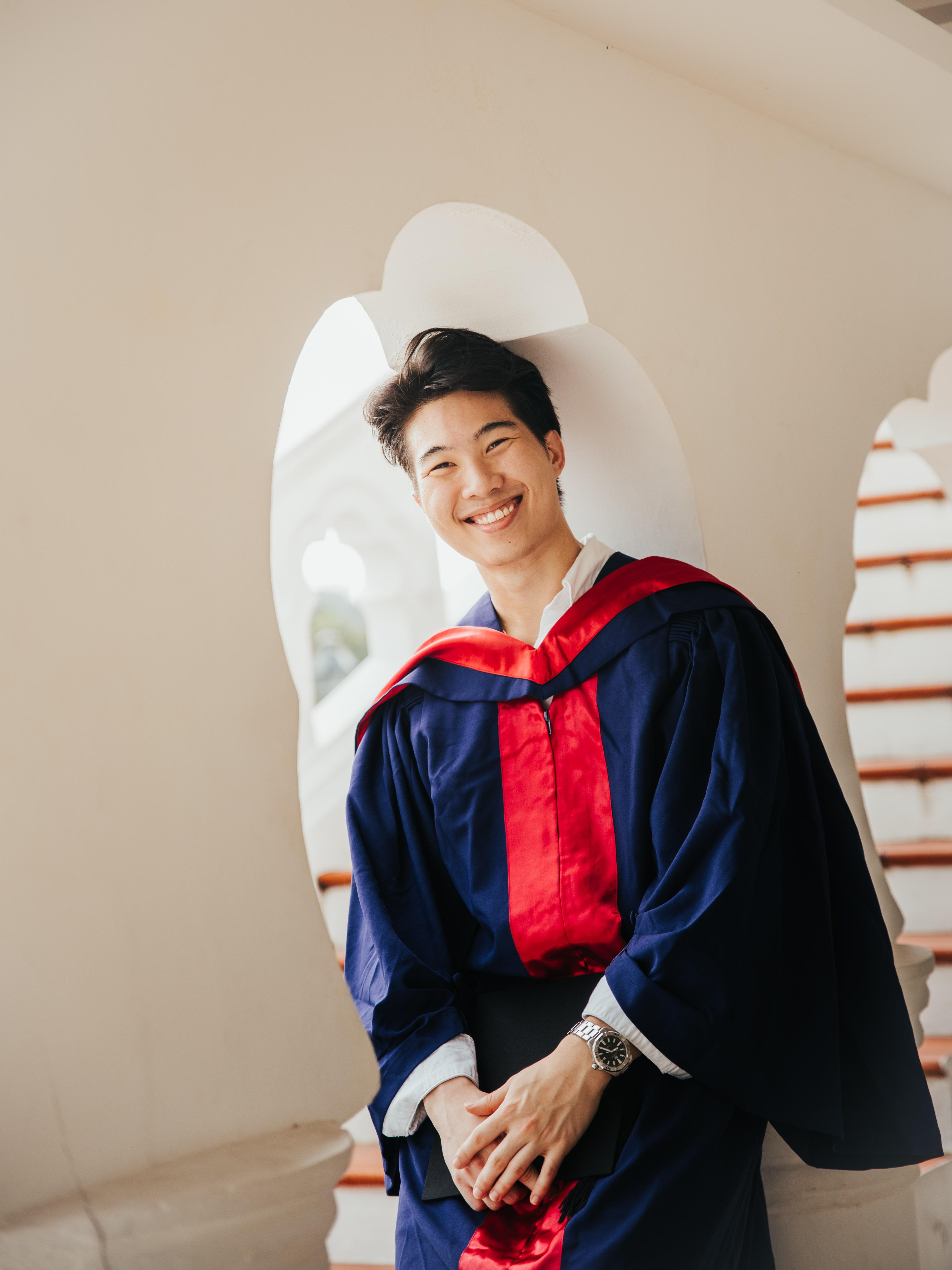 A person wearing a graduation gown and cap smiles while standing indoors near a staircase.
