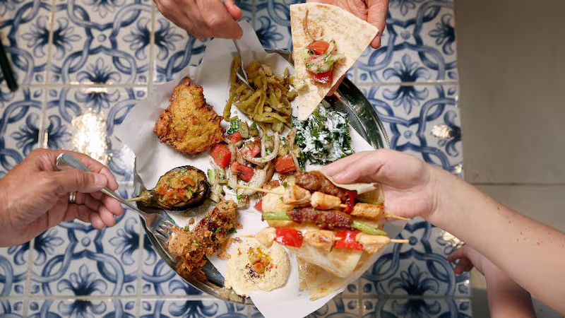Wooden outdoor table set with multiple plates of food, including dips, salads, potatoes with cheese, and glasses of water, with hands reaching in to eat.