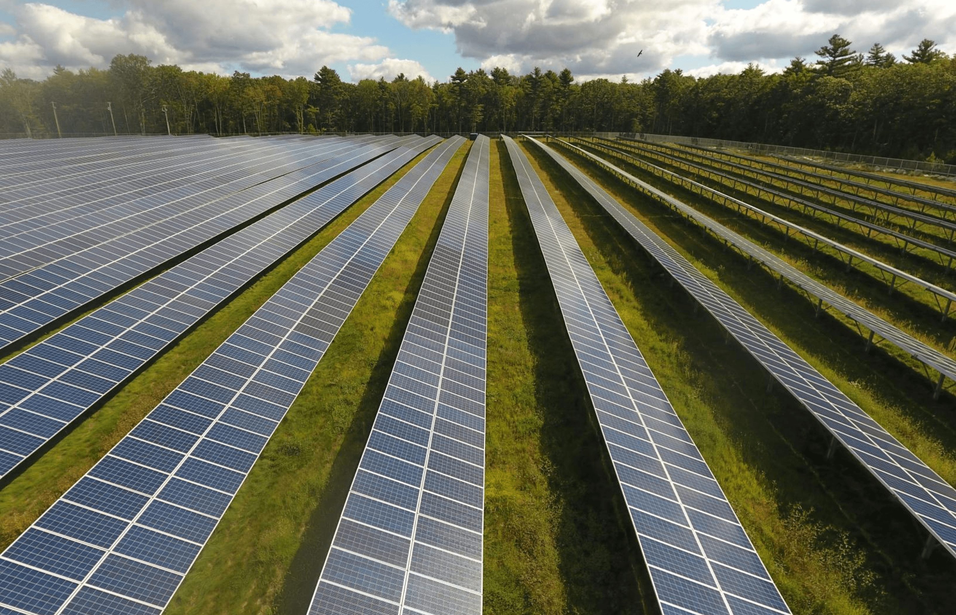 Aerial view of solar panels arranged in rows on a green field under a blue sky with scattered clouds.