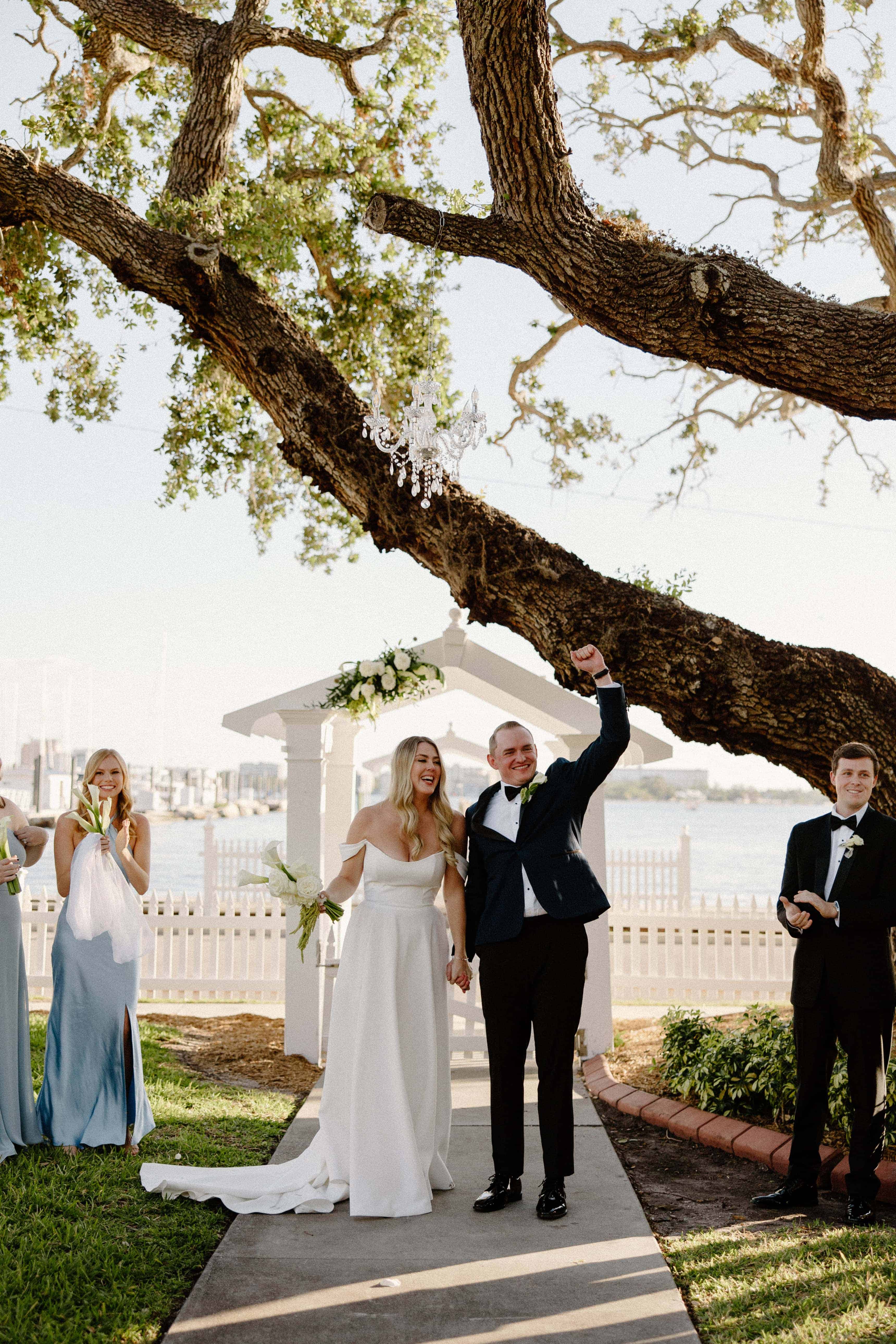A photo of a wedding couple standing under a tree with guests in the background. The groom is raising his arm in the air.