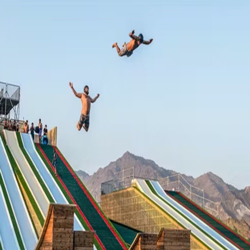 رجلان في الهواء بعد التزحلق على منزلقات مائية ملونة، مع الجبال في الخلفية والمتفرجين على الجانبين.