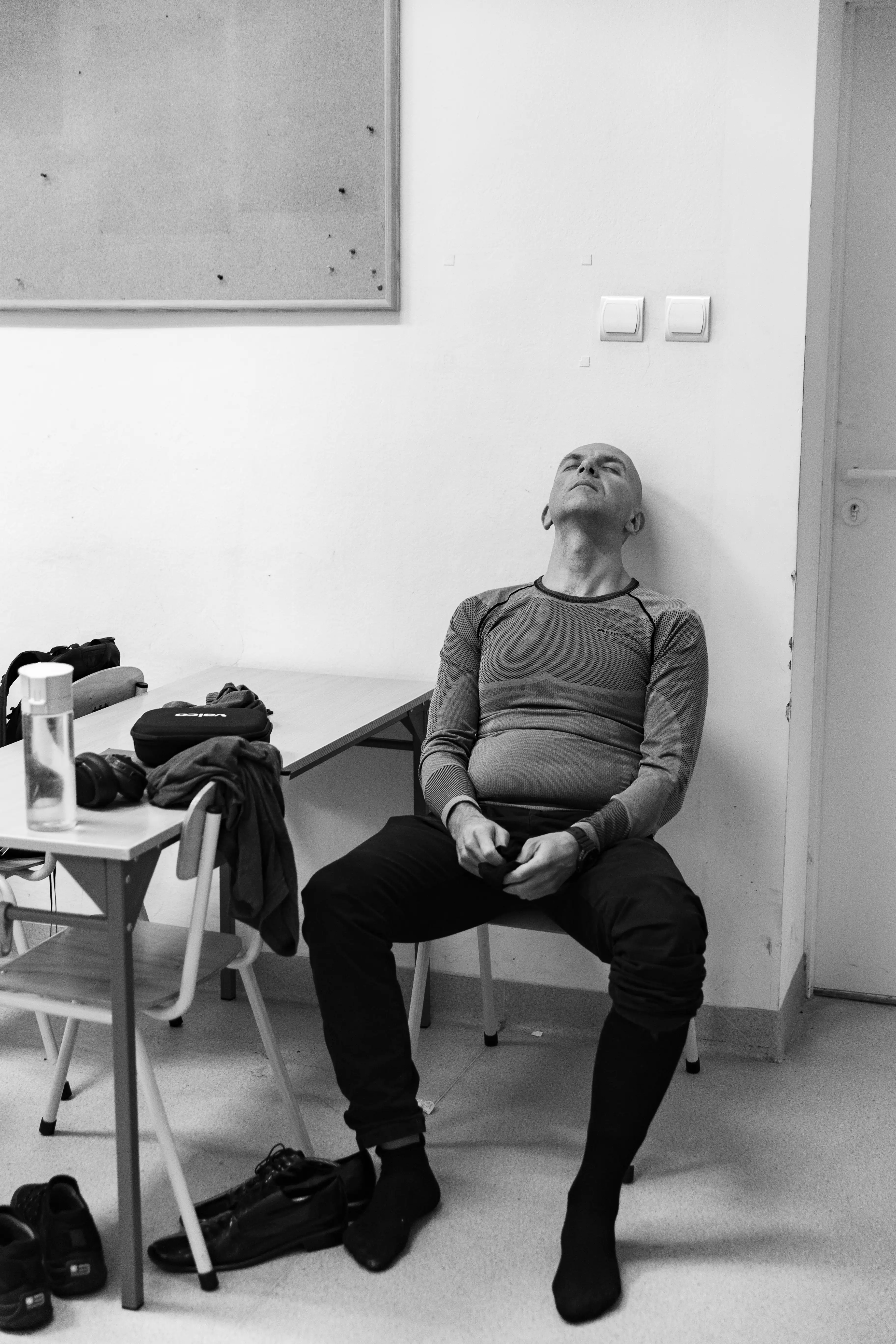 Mężczyzna w sportowym stroju siedzi na krześle, oparty o ścianę. Ma zamknięte oczy i ręce na kolanach. W tle widać nieuporządkowane rzeczy oraz część pomieszczenia.