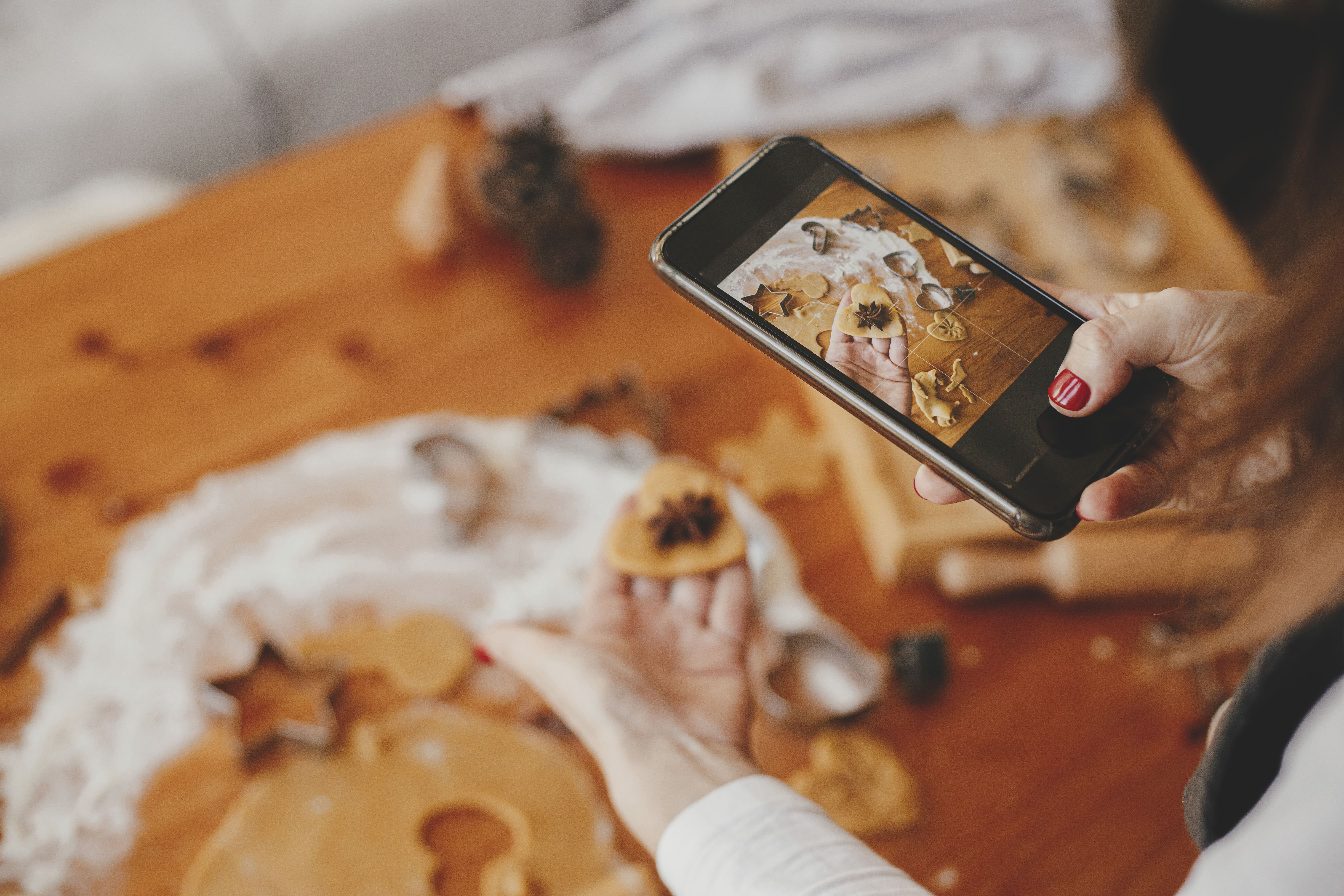 Woman photographing handmade gingerbread cookies with a smartphone during a baking session at home.