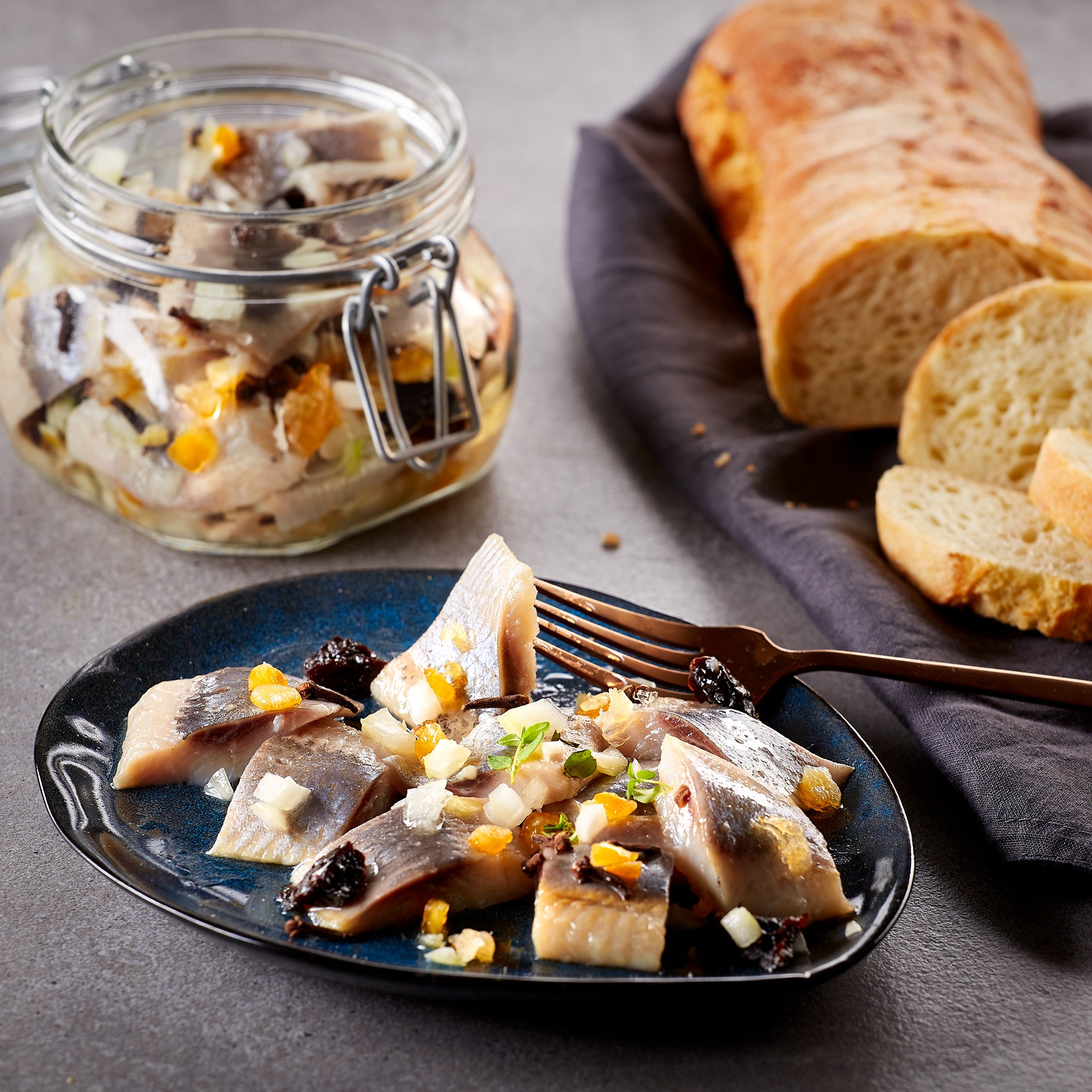 A rustic dining scene features marinated herring pieces garnished with herbs and diced vegetables on a dark plate, complemented by a loaf of crusty bread and a jar of additional marinated fish, creating an inviting appetizer presentation. 