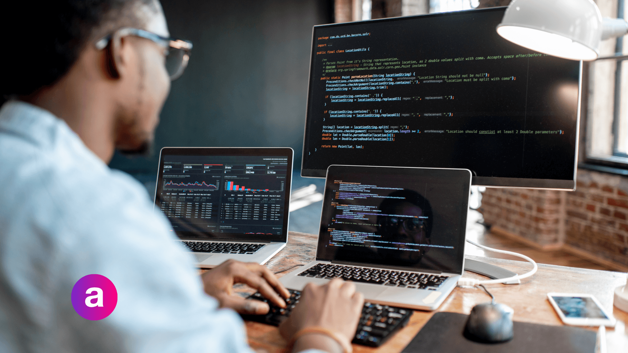 Person wearing glasses working as a software engineering with two laptops and a monitor on a desk in front of them
