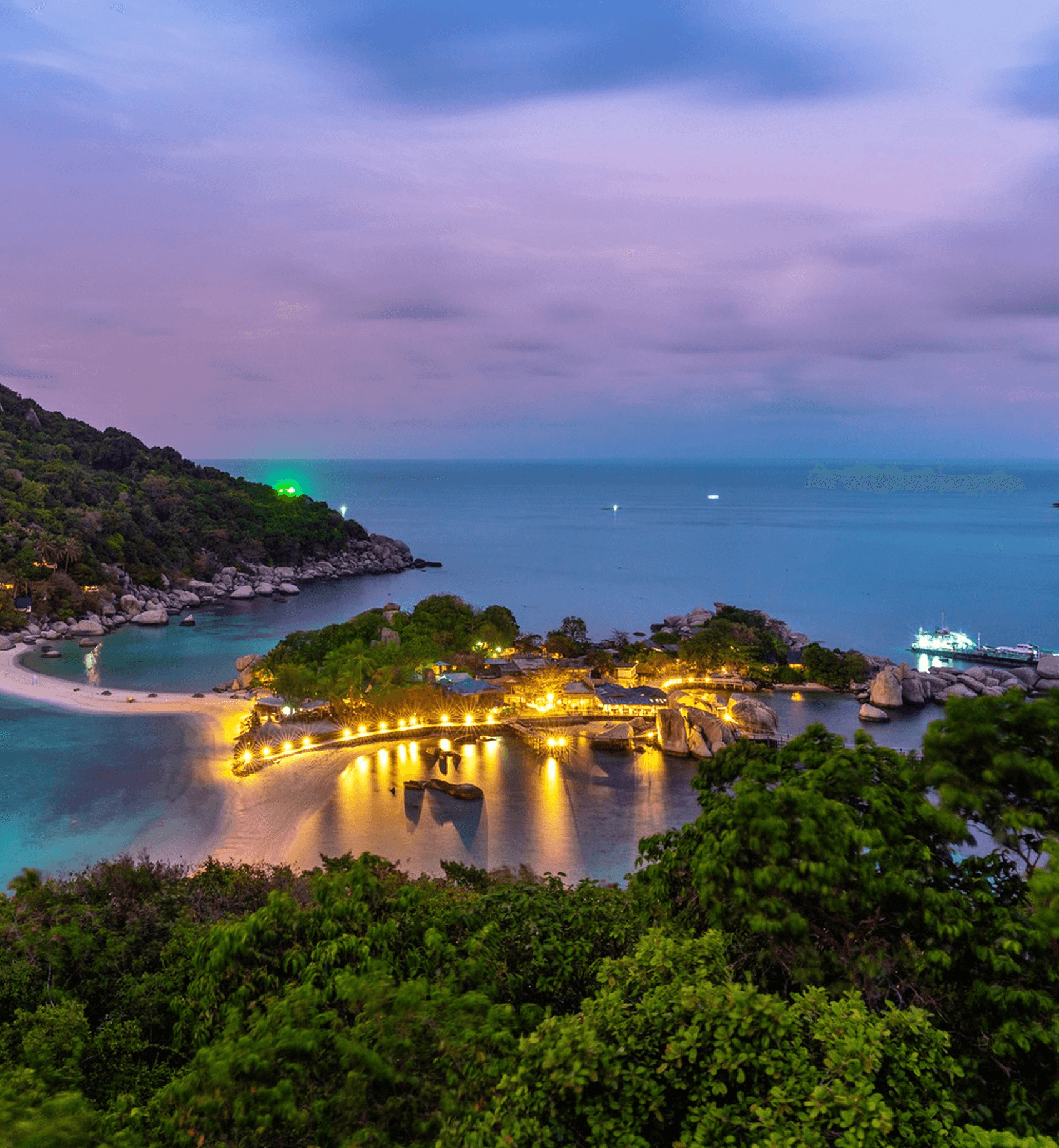 Tropical island bay at dusk with glowing shoreline lights and calm blue water.