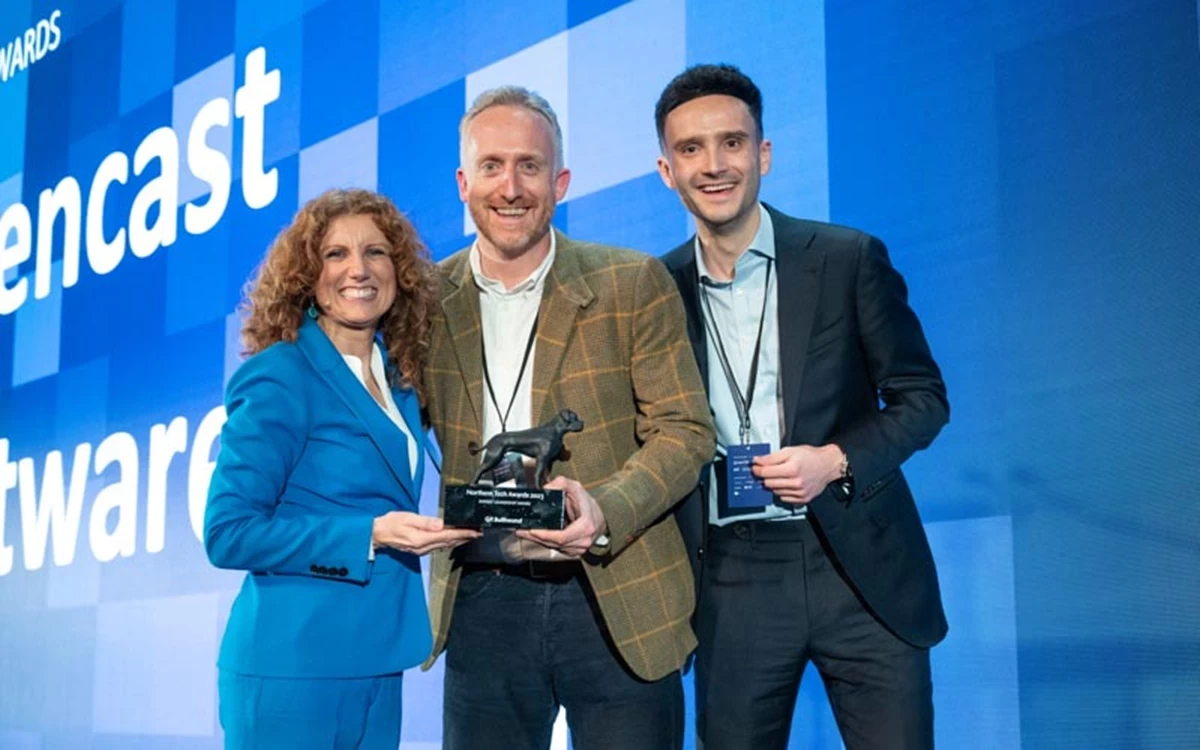 Three people stand on stage holding an award trophy in front of a large screen with event branding. They are dressed in business attire and smiling toward the camera.