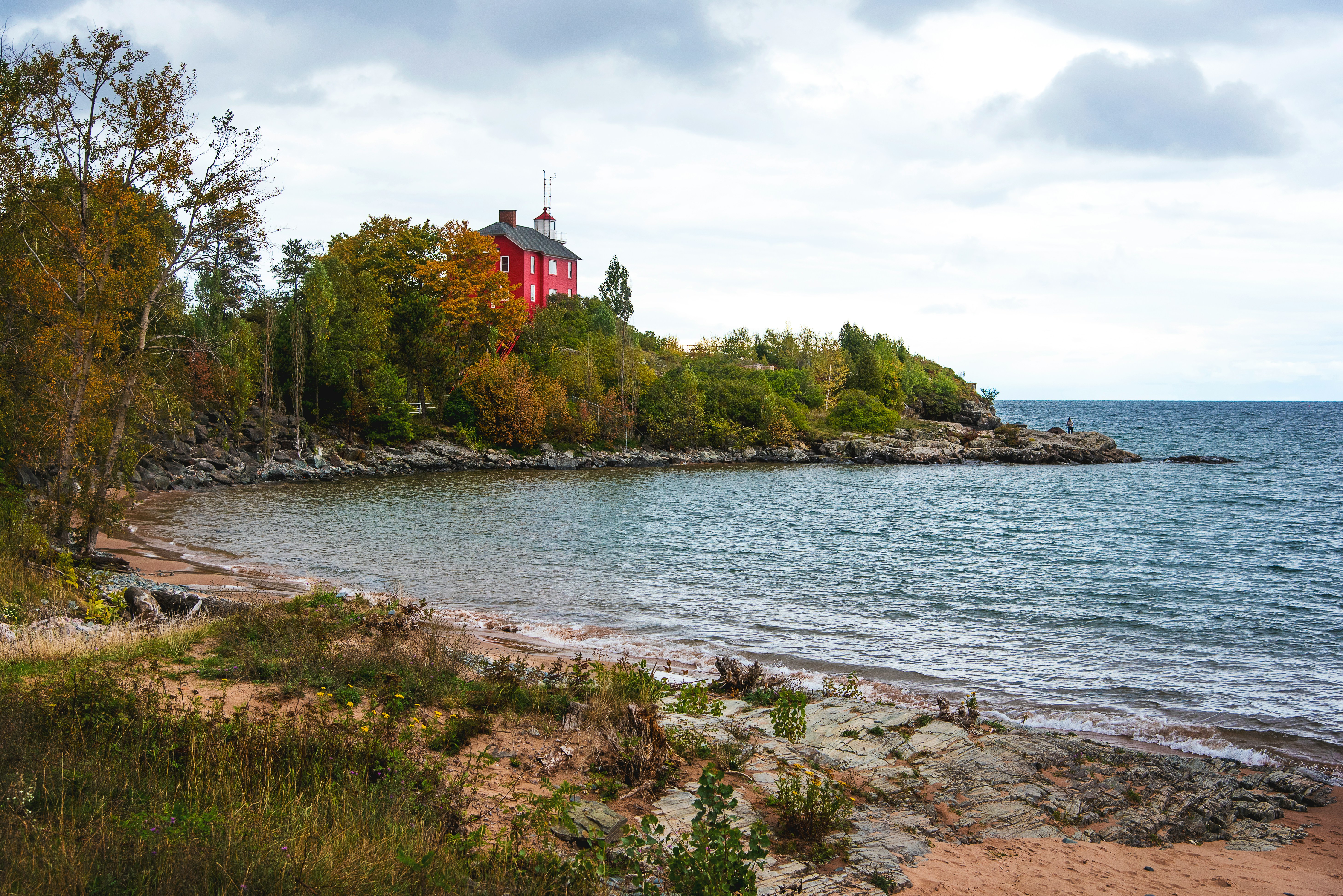 A serene shoreline with rocky beach, lush greenery, and a distant house against a cloudy sky.