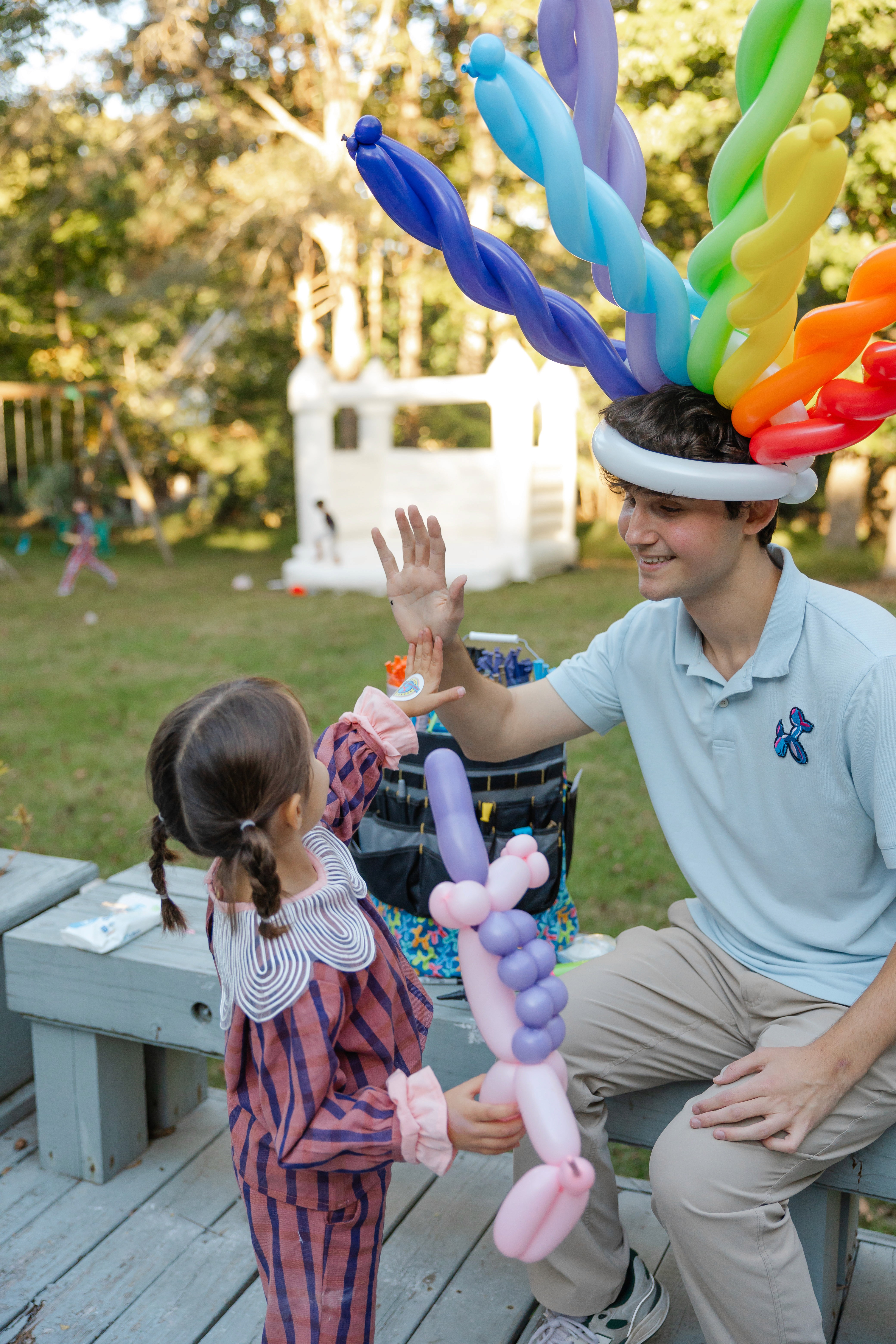 A man in a light blue polo shirt wearing a colorful balloon hat sits on an outdoor deck, giving a high-five to a young girl holding a pink balloon unicorn, with a playhouse and greenery visible in the background