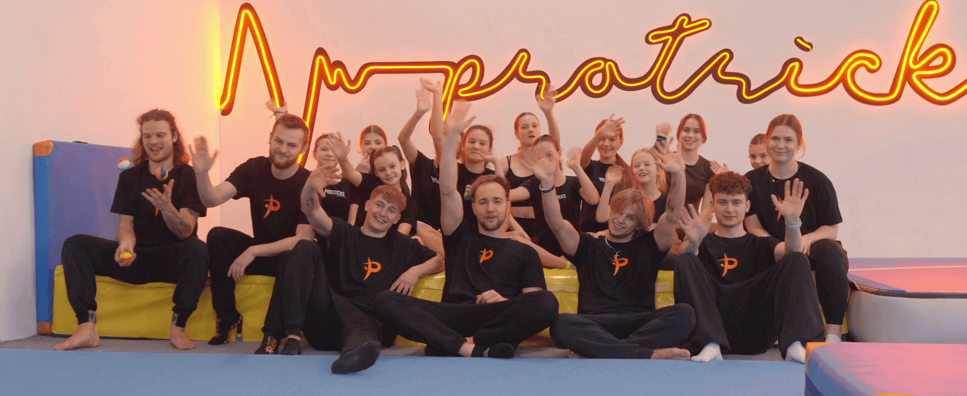 Grupa trenerów i uczniów zajęć z gimnastyki entuzjastycznie machają w sali gimnastycznej na tle neonowego napisu "ProTricks"