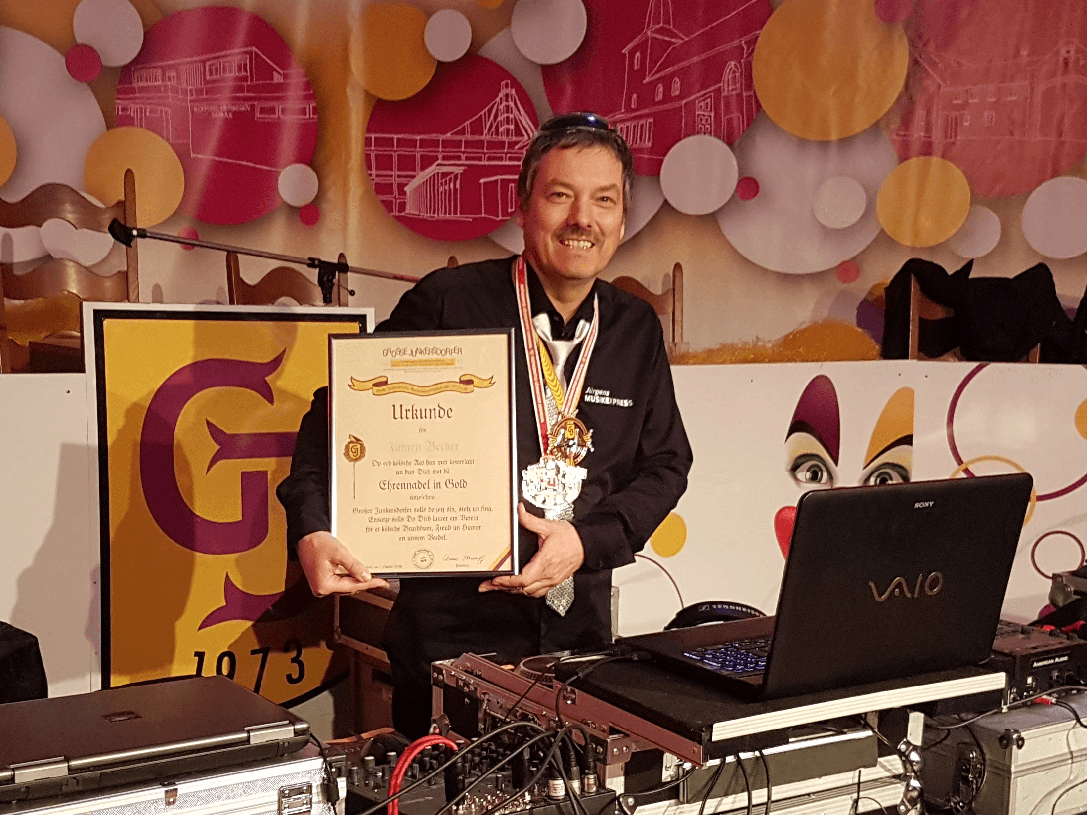 Ein Mann vor DJ Pult mit Auszeichnung in den Händen. Freundlich, symphatisch. Karnevalistische Kulisse