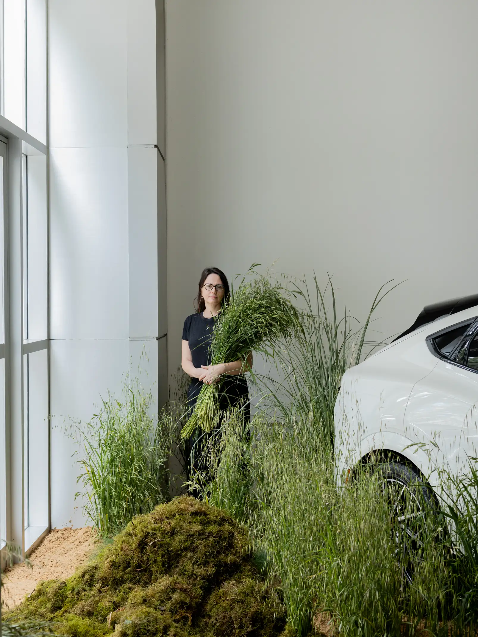 Irina Khatsernova sosteniendo follaje verde en instalación botánica contemporánea junto a un automóvil blanco en espacio minimalista e iluminado.
