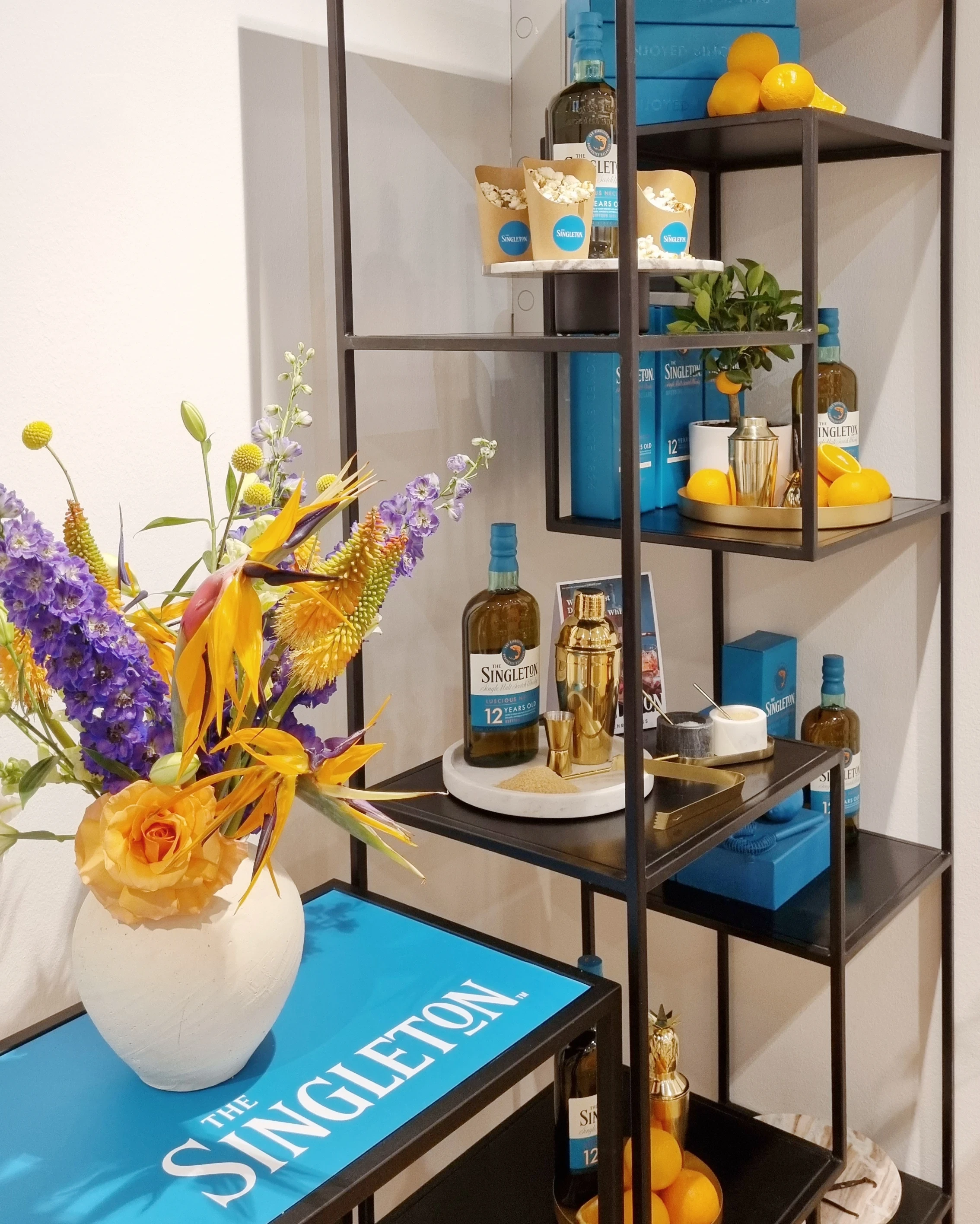 Black metal shelf with whisky bottle, golden shaker, snacks and lemons next to a colourful bouquet.