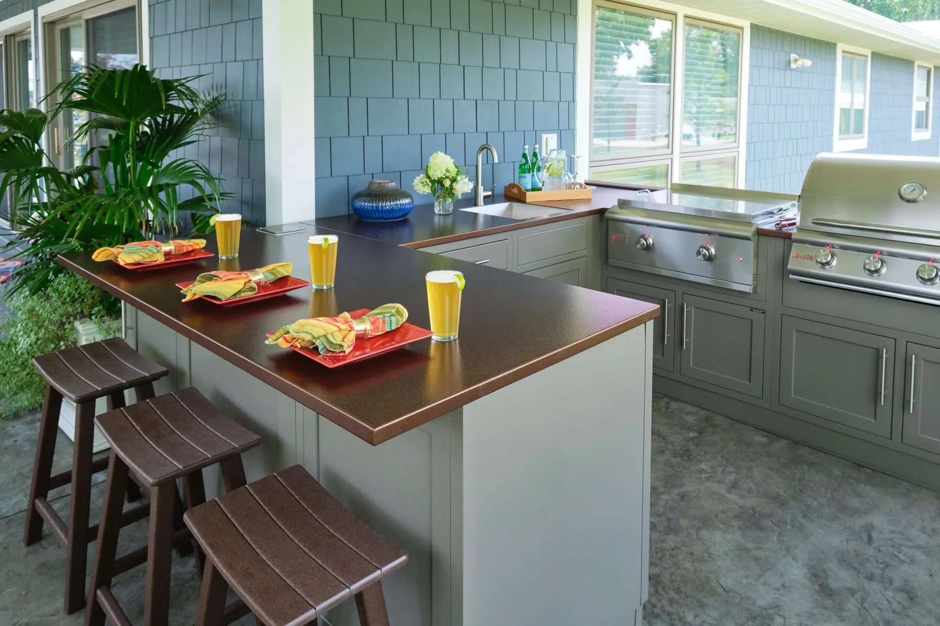 An outdoor kitchen with gray cabinets, a built-in grill, and a contrasting dark countertop that extends into a bar area with wooden stools. Brightly colored plates, napkins, and drinks decorate the bar, with lush plants and a cozy setting completing the space.