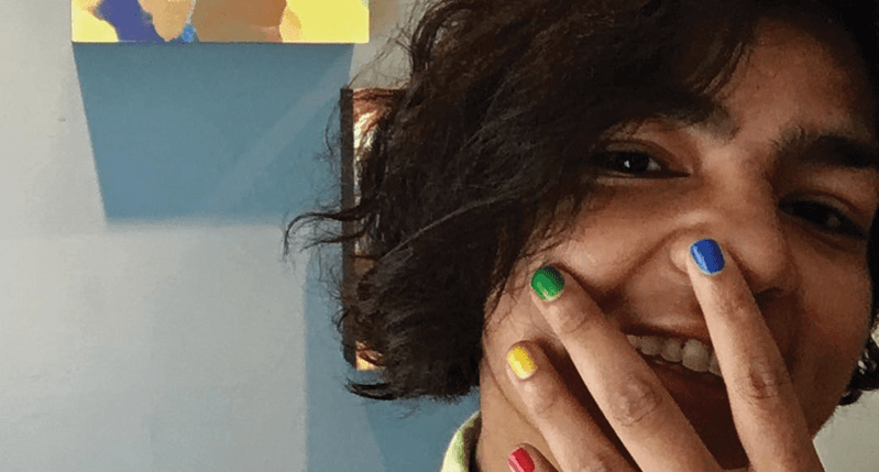 A photo of a woman with her hands over her face. Her nails are colored in yellow, red, blue and green.