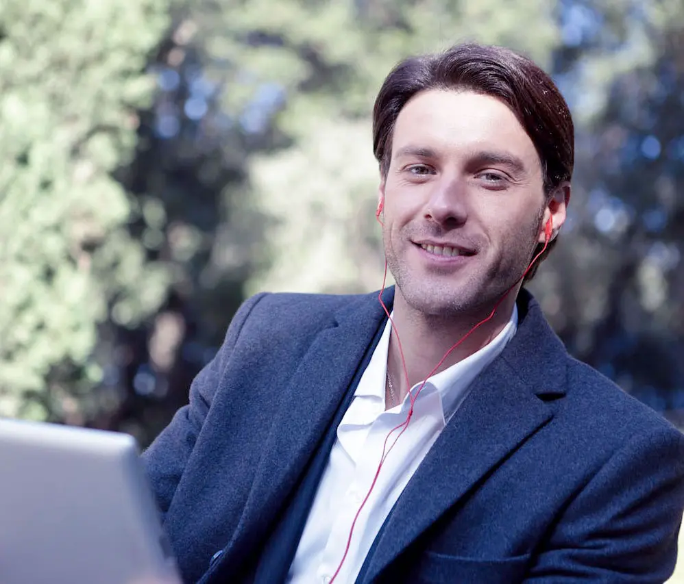 Man in suit wearing earbuds, smiling and holding a laptop outdoors.