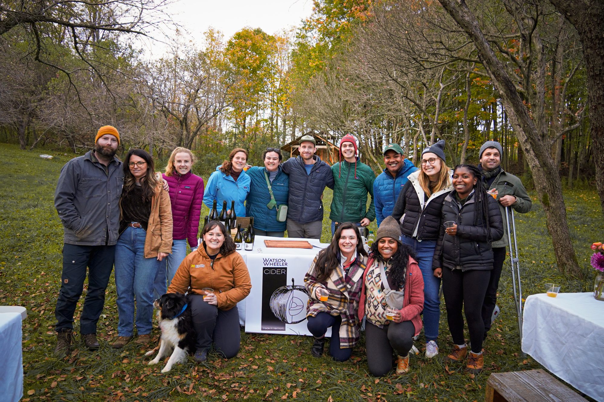 A diverse group of people stands together outdoors, smiling, with trees and nature in the background.