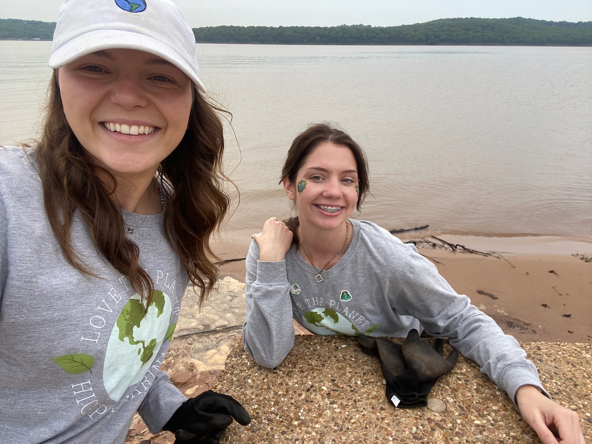 Two people wearing matching earth-themed shirts and hats with globe logos, smiling at the camera while sitting on a sandy lake shoreline, with forested hills in the background.