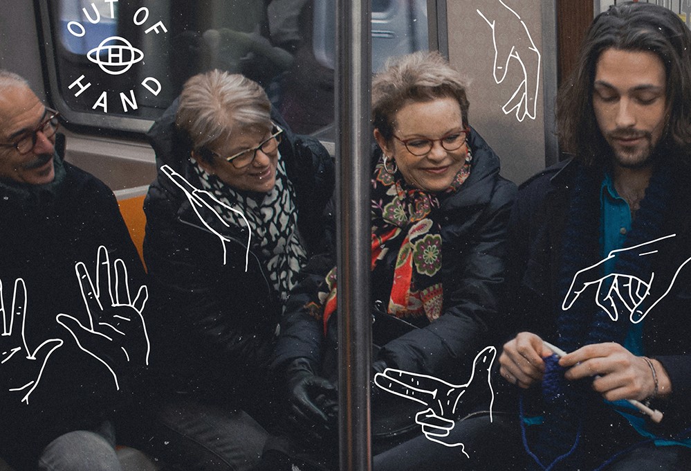 Three people are seated on a subway train. A woman with short gray hair and glasses sits next to a man with long dark hair and a beard. A third person with short light hair and glasses is seated between them. The subway car window displays white line drawings of hands and the words “OUT OF HAND” above the window.