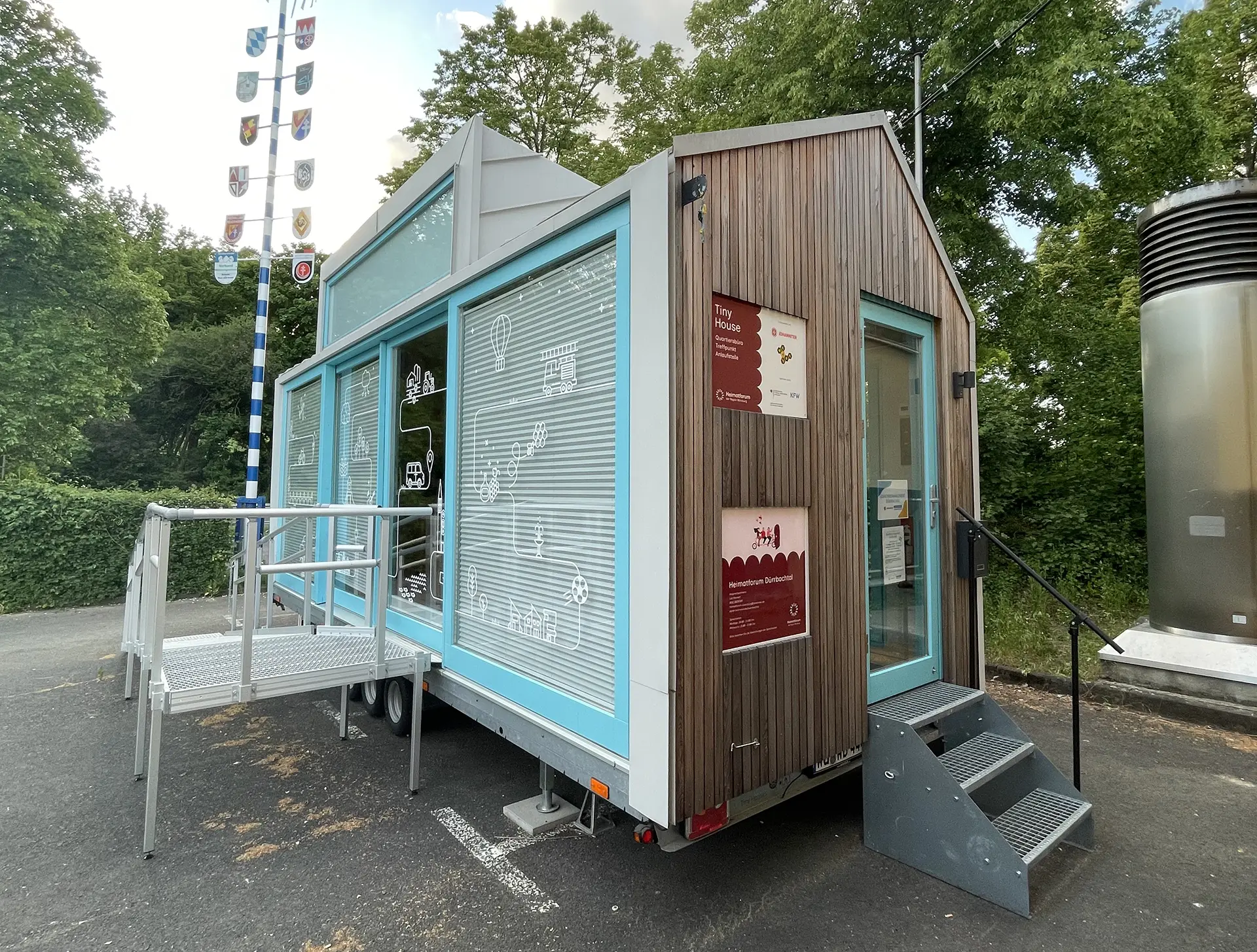 Tiny Houses auf Anhänger mit türkisfarbenen Fenstern, einer Holzfassade, dekorativen Piktogrammen und Informationsschildern. Ausgestellt auf einem Außengelände mit Bäumen im Hintergrund.