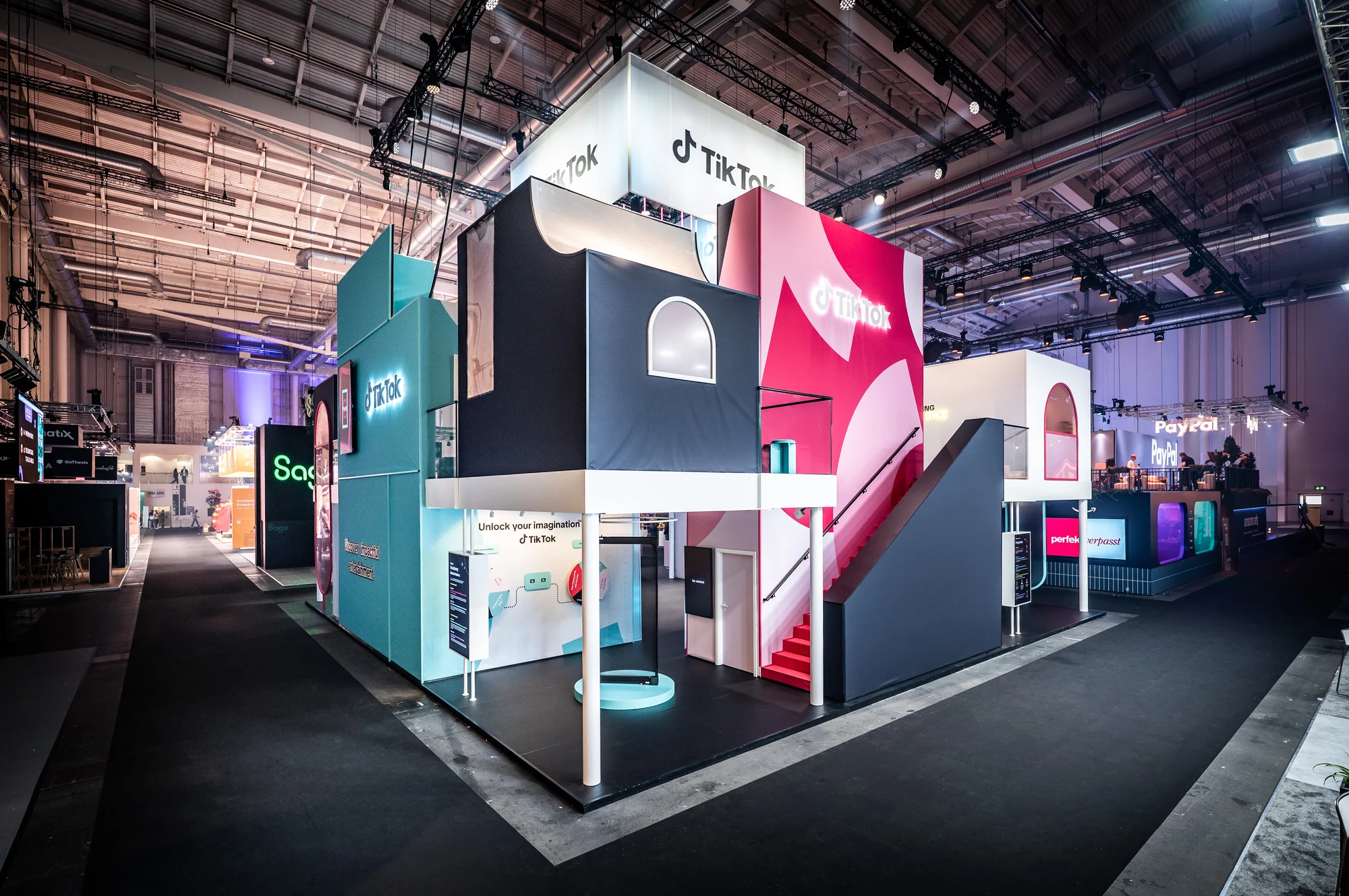 Exterior view of the multi-storey TikTok exhibition booth, combining navy and pink modular facades with branded signage and stair access.