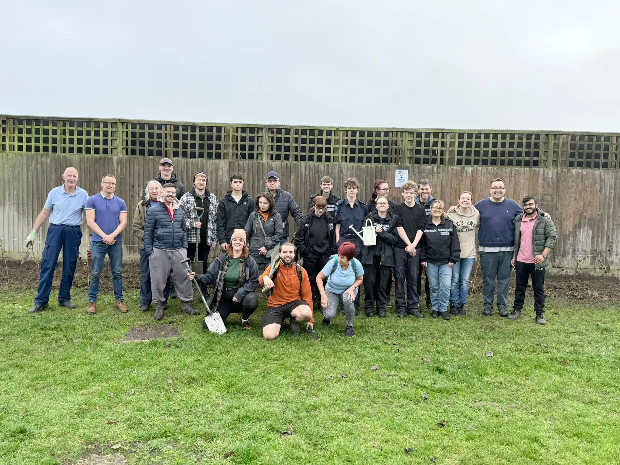 A group of people standing together outdoors, smiling, with green grass and a stone wall in the background.