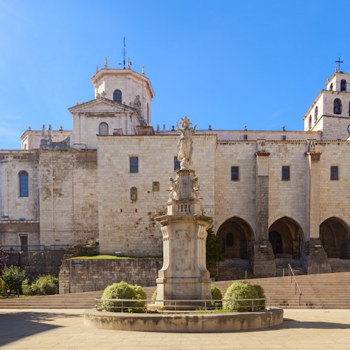 Large stone cathedral with arched entrances, a central fountain with a statue, and manicured bushes, under a clear blue sky.