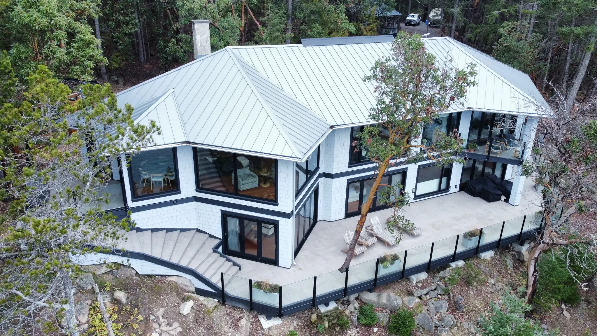 Aerial view of a modern hillside home with a large wraparound deck and outdoor living space surrounded by forest.