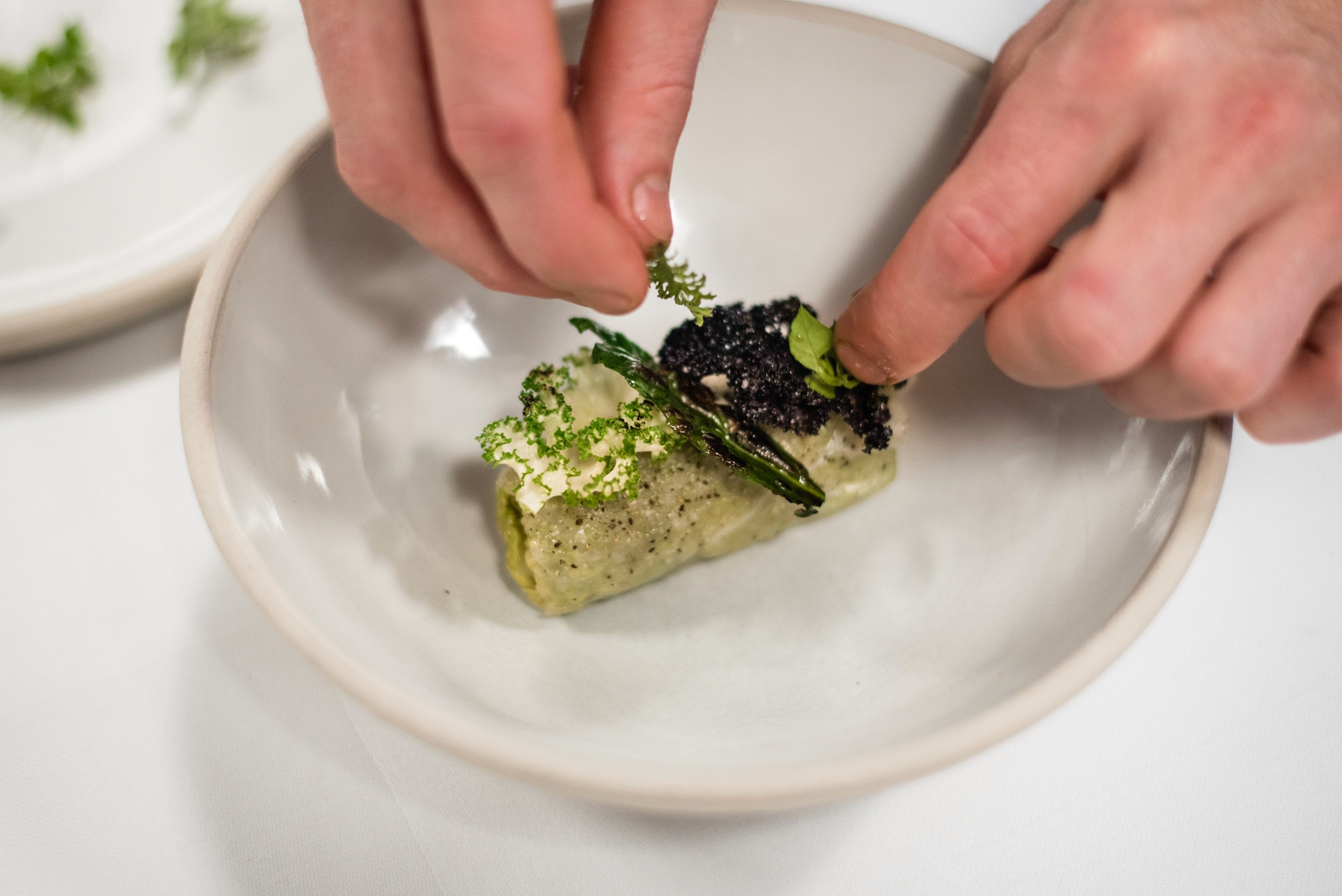 A chef's hands garnish a cylindrical piece of seared meat topped with black caviar and green herbs on a white plate.