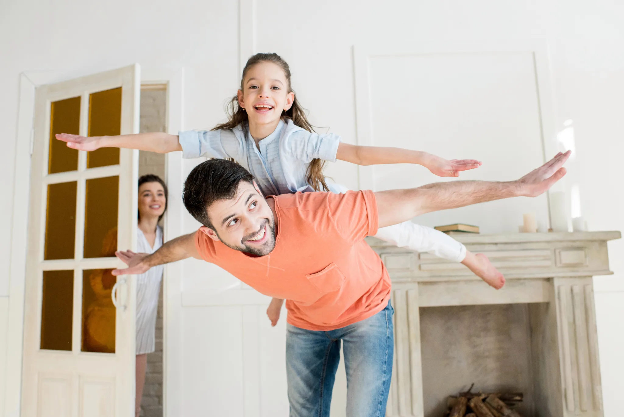 Daughter on her dad's back, both have their arms extended like they are flying, as mom smiles behind them in the doorway.