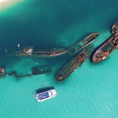 Aerial view of a white boat near multiple submerged shipwrecks in clear, turquoise water.