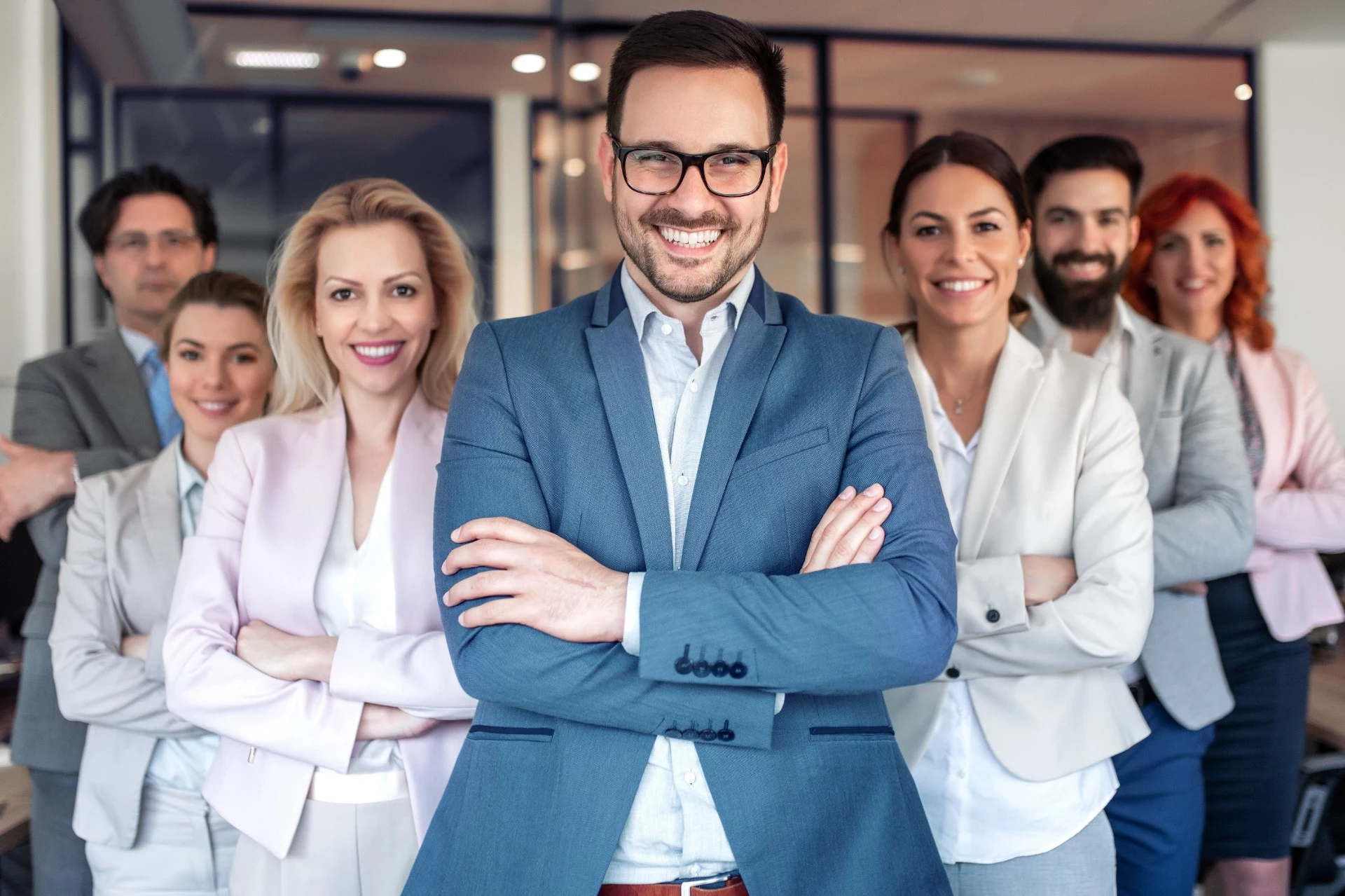 Imagem de um grupo de profissionais em um ambiente corporativo, com um homem sorridente e de braços cruzados em destaque no centro, vestindo um terno azul e óculos. Ao fundo, uma equipe diversificada, todos com expressões amigáveis e postura confiante, transmite uma imagem de união e profissionalismo. O fundo, com janelas de vidro e portas, sugere um escritório moderno e iluminado.