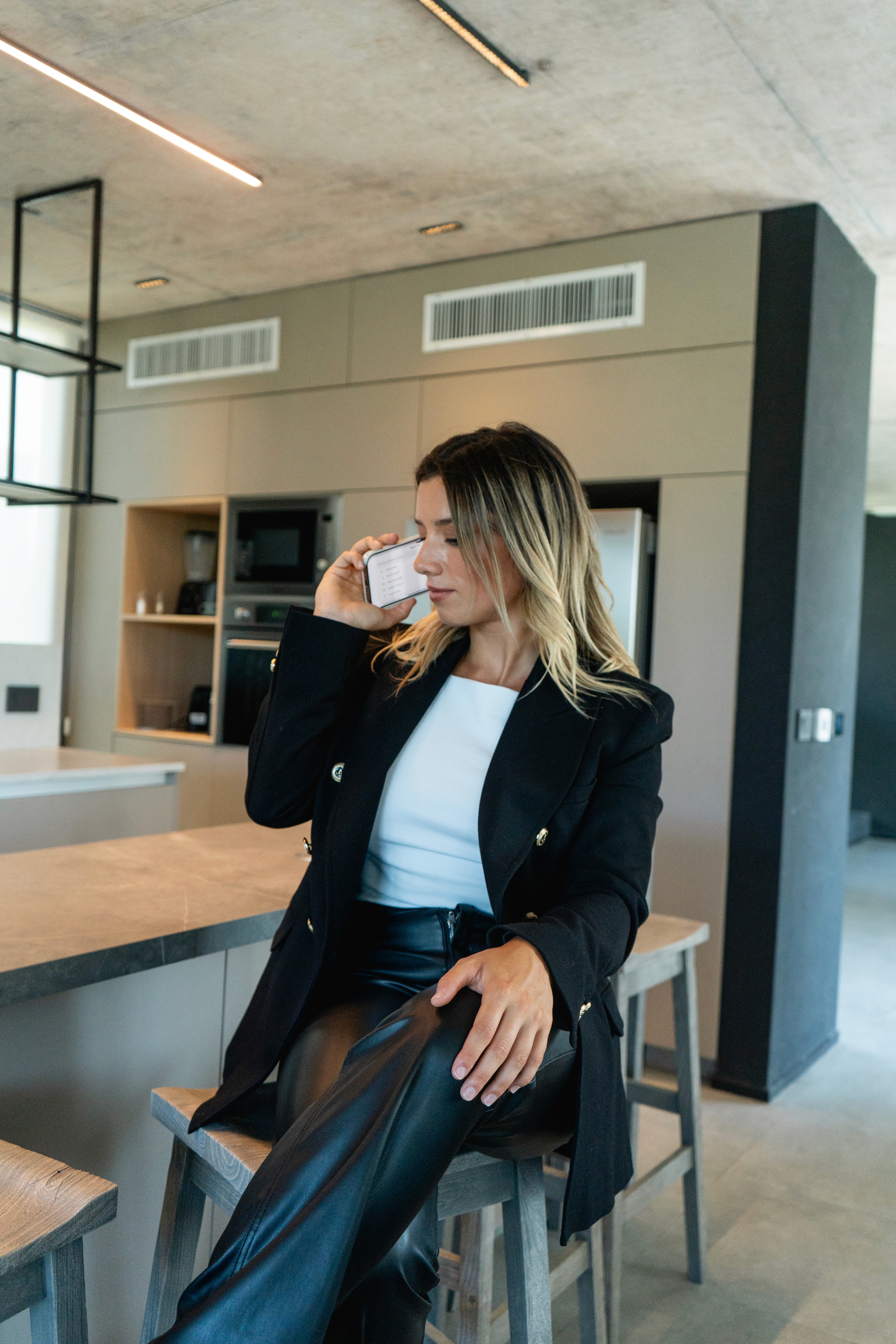Mujer con blazer negro y pantalón de cuero, sentada en banqueta de cocina moderna, habla por teléfono móvil.