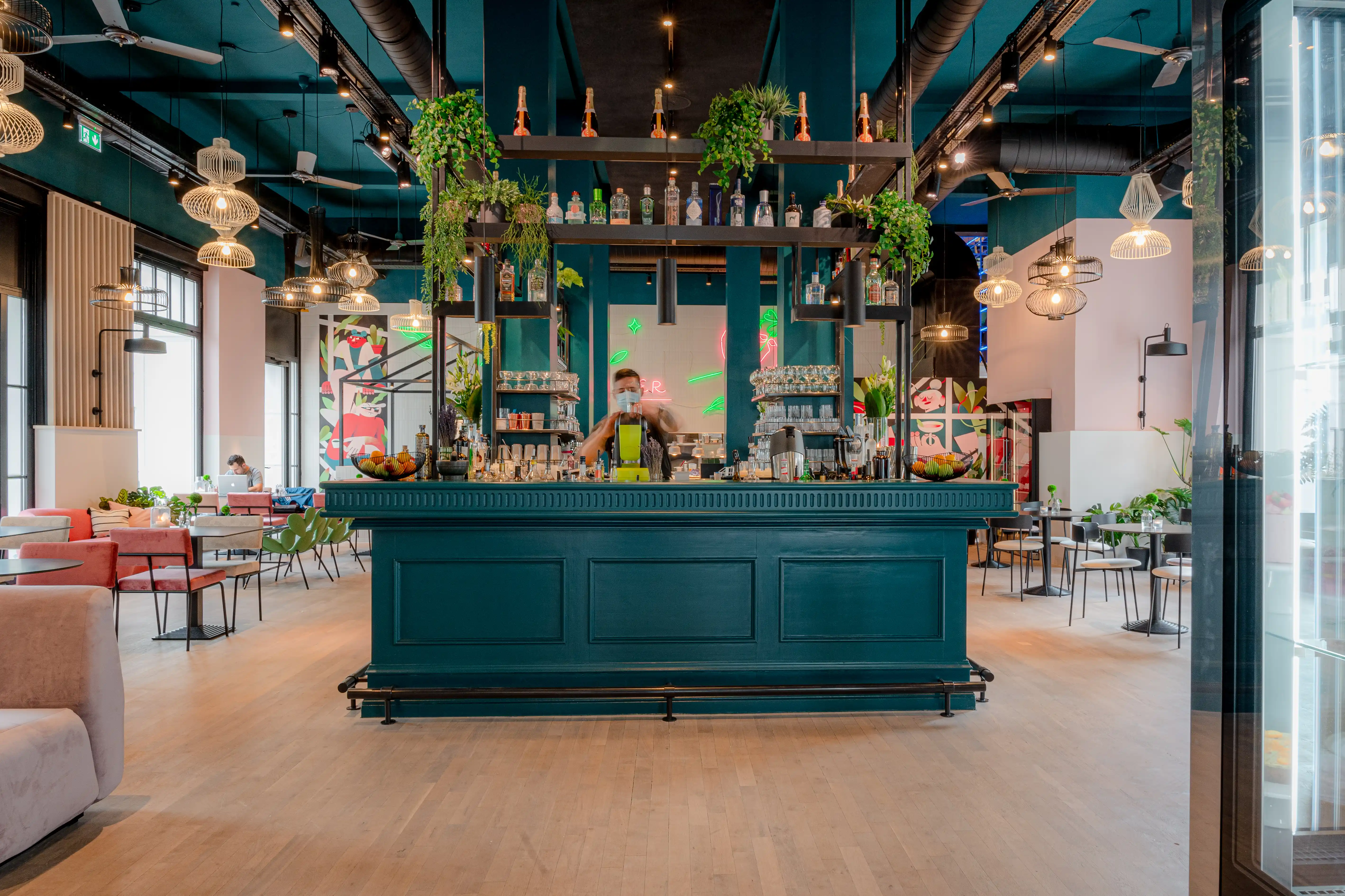 Central teal bar at LOKAL VIER with classic panelled front, hanging plants on a black steel rack, bottle shelves and a barista working between wire pendant lights.