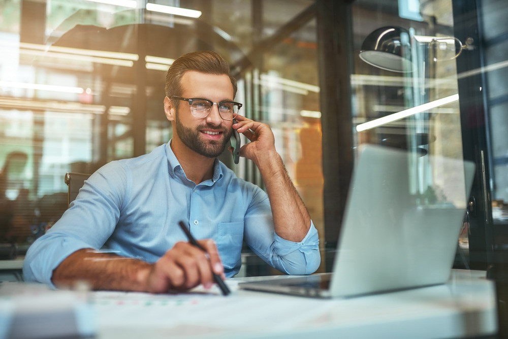 Customer support. Portrait of young and cheerful businessman in eyeglasses and formal wear talking on the phone and smiling while sitting in the office