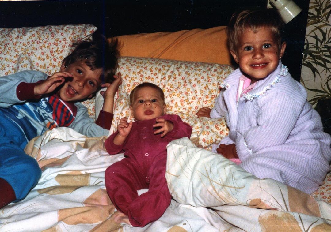 Childhood photo of the Korman siblings: Three young children lying on a bed, smiling and posing for the camera.