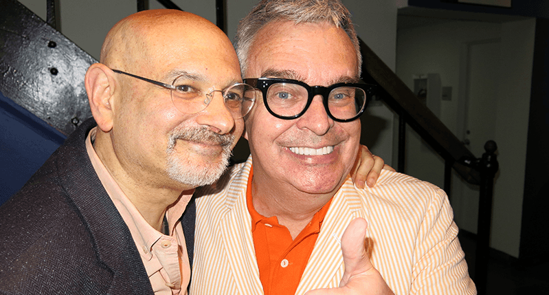 A photo of two men, wearing glasses, standing in an exhibition room.