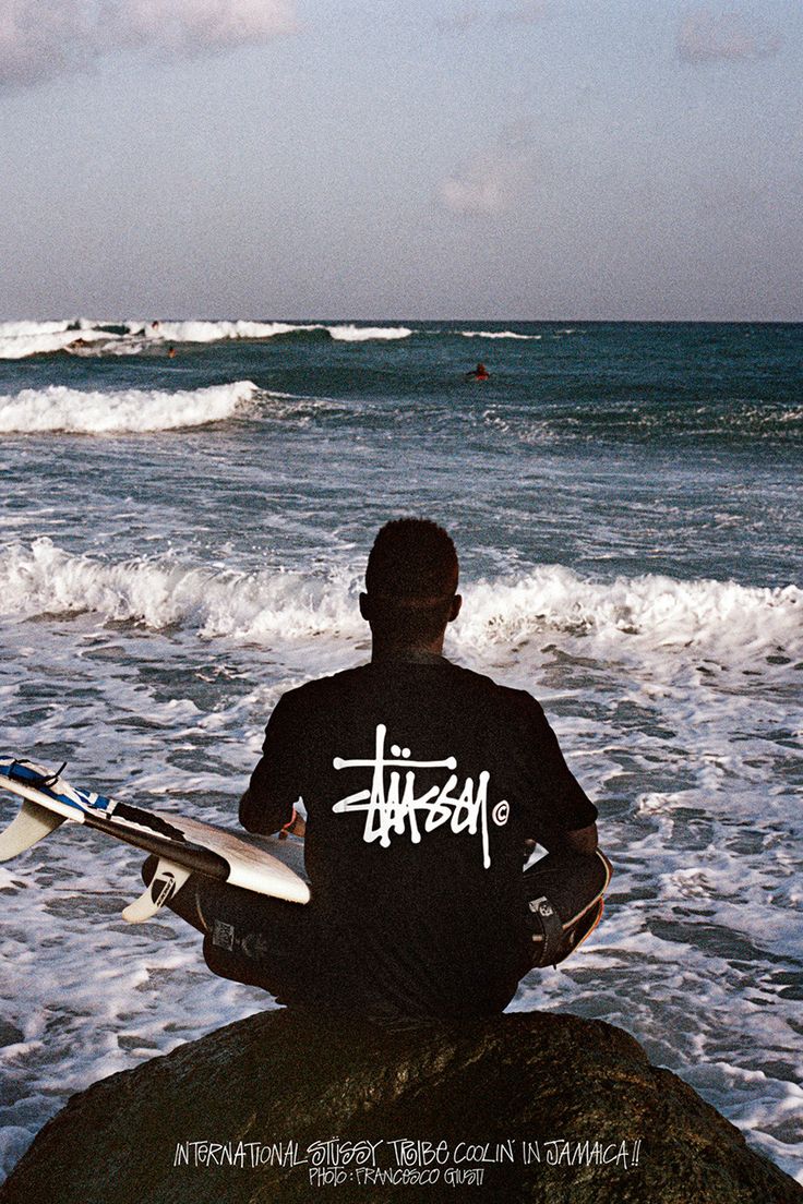 a stussy ad showing a man sitting on a rock looking out at the waves while holding a surf board