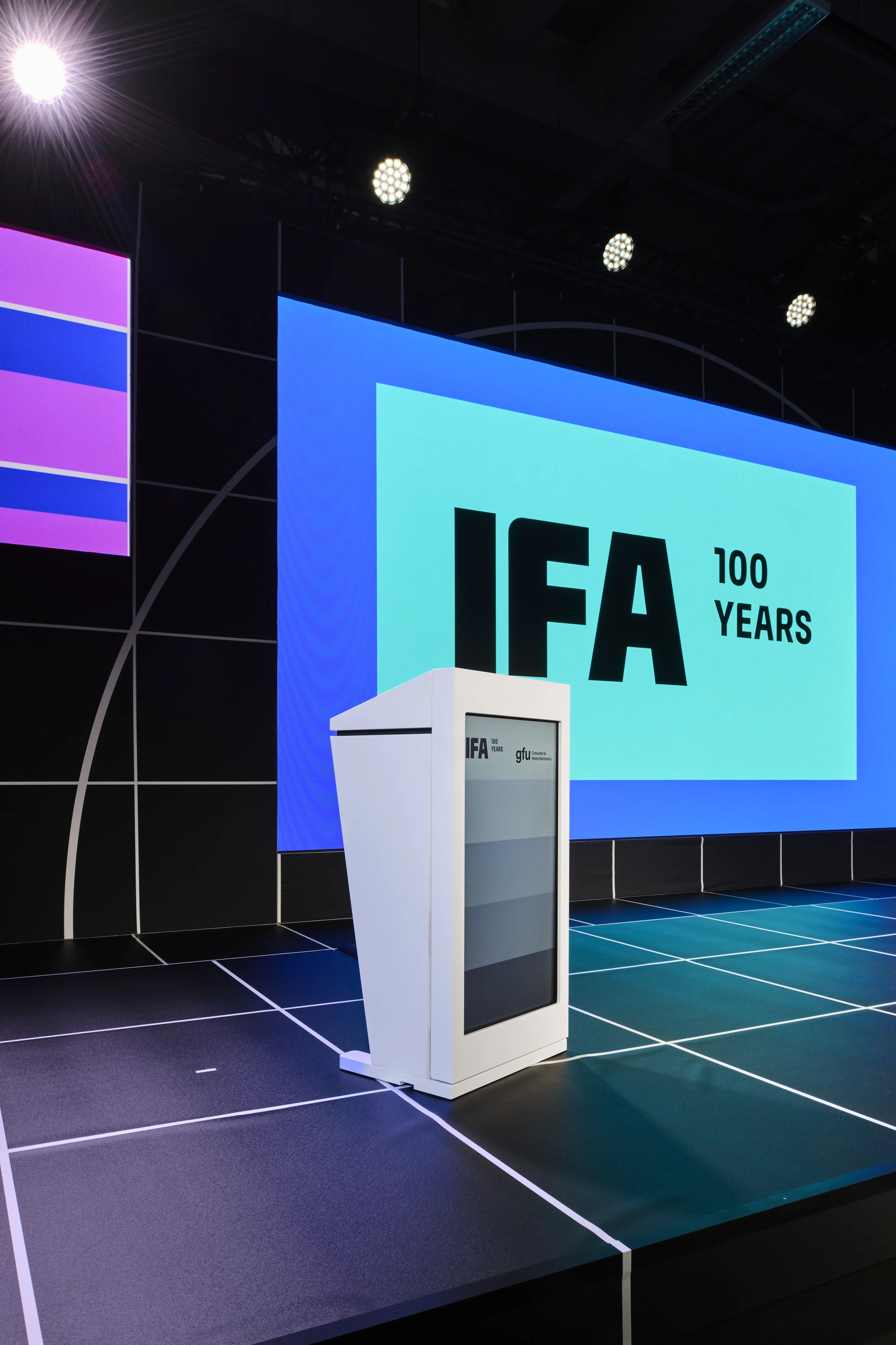 White lectern on the black grid stage in front of a turquoise IFA 100 Years screen.