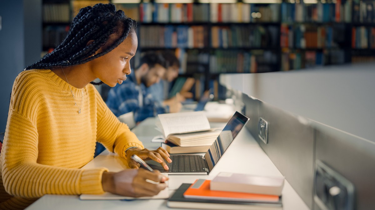 Young woman sitting in a library, reading on her computer and taking notes 