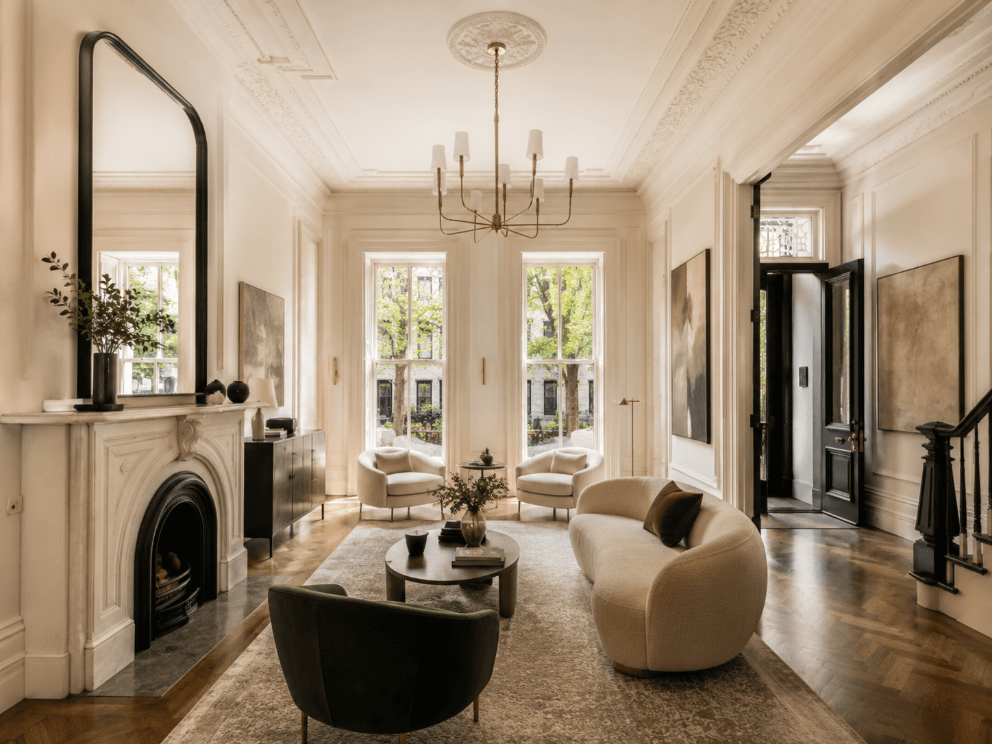 Elegant parlor-level living room inside a renovated New York brownstone, featuring tall windows, ornate ceiling moldings, a fireplace, herringbone wood floors, and luxury neutral furnishings.