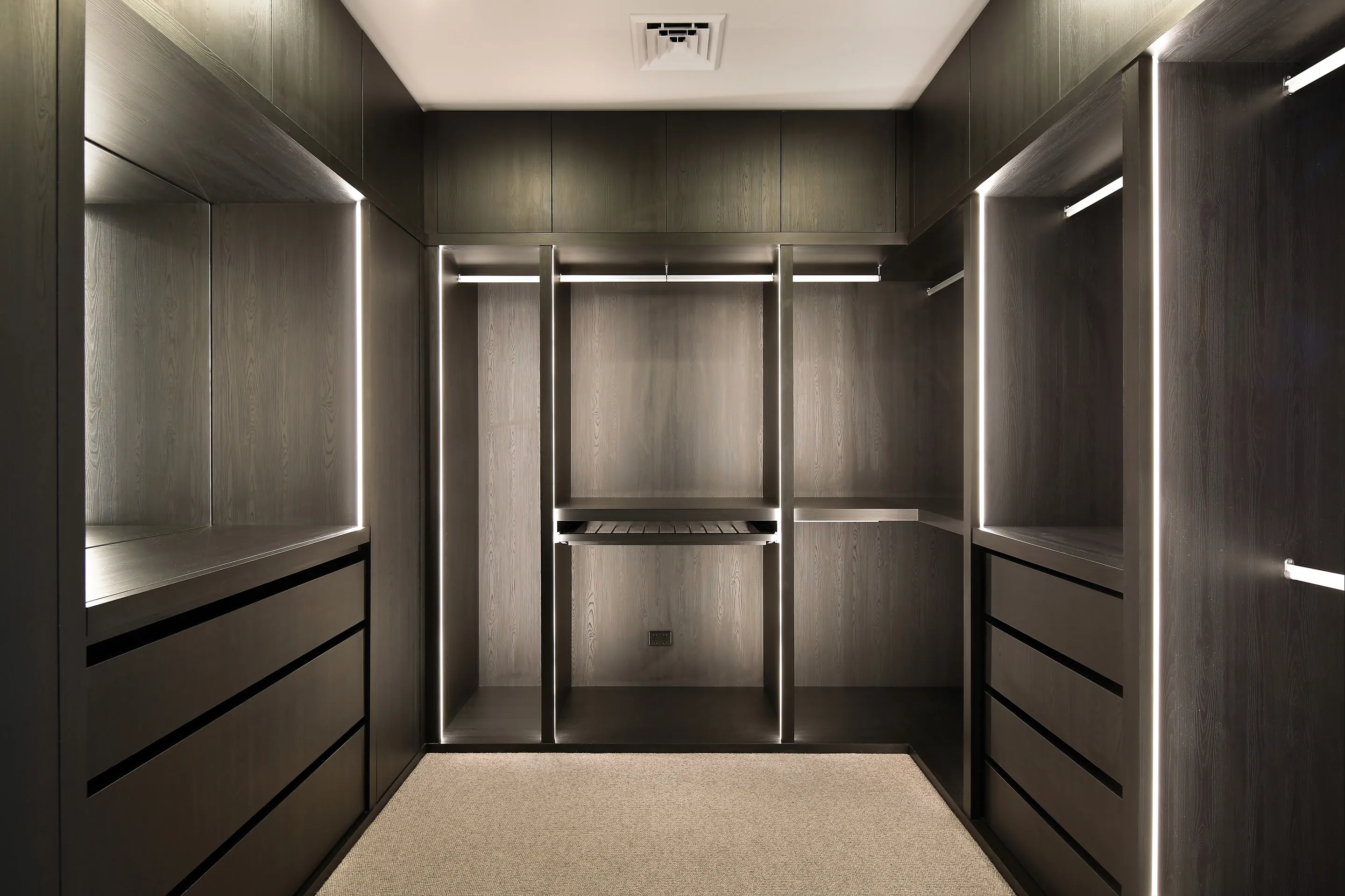 Walk-in robe with dark timber-veneer joinery, open shelving, hanging rails with integrated LED strips, banks of drawers and a pull-out shoe rack over beige carpet