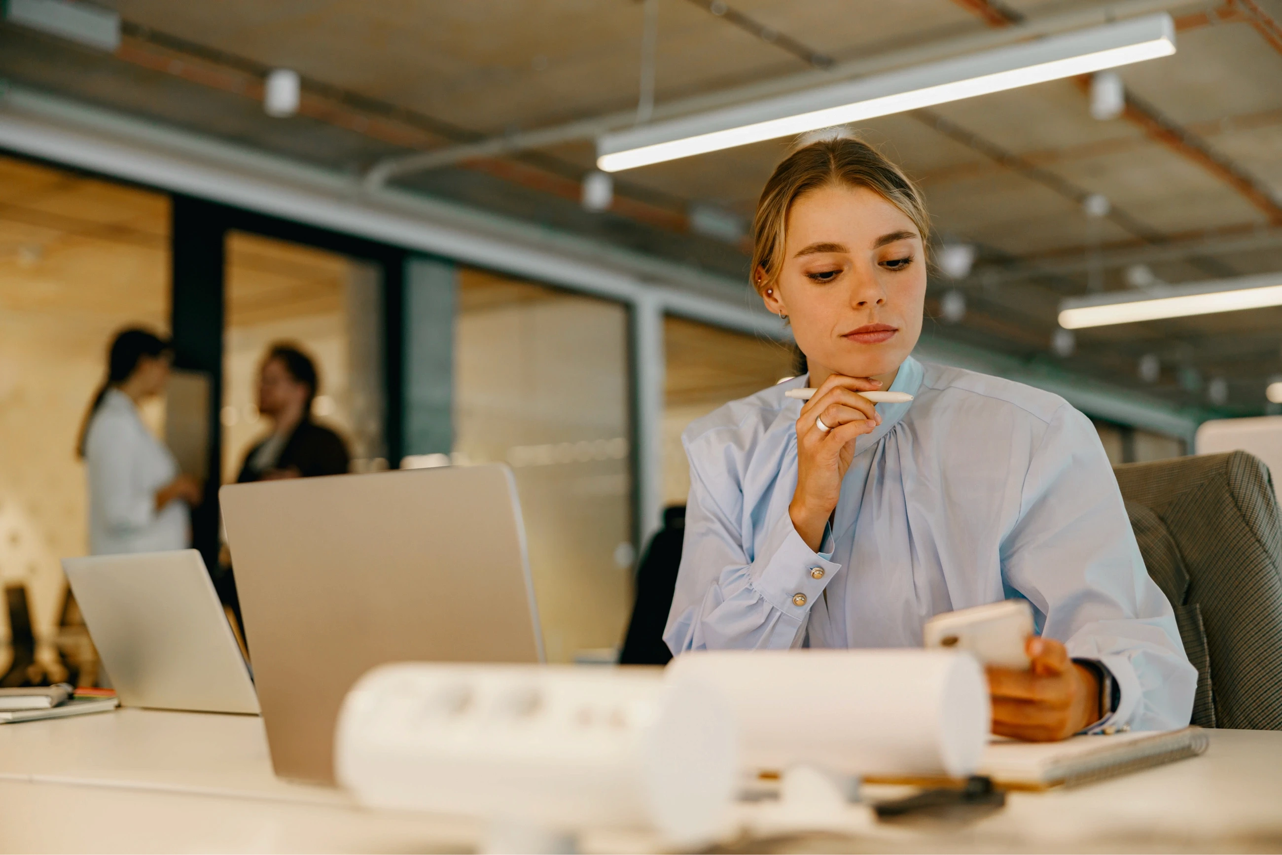 In a modern office space, a person in a blue shirt with a focused expression sits at a white desk, holding a smartphone and pen, in front of a laptop with blurred colleagues in the background.
