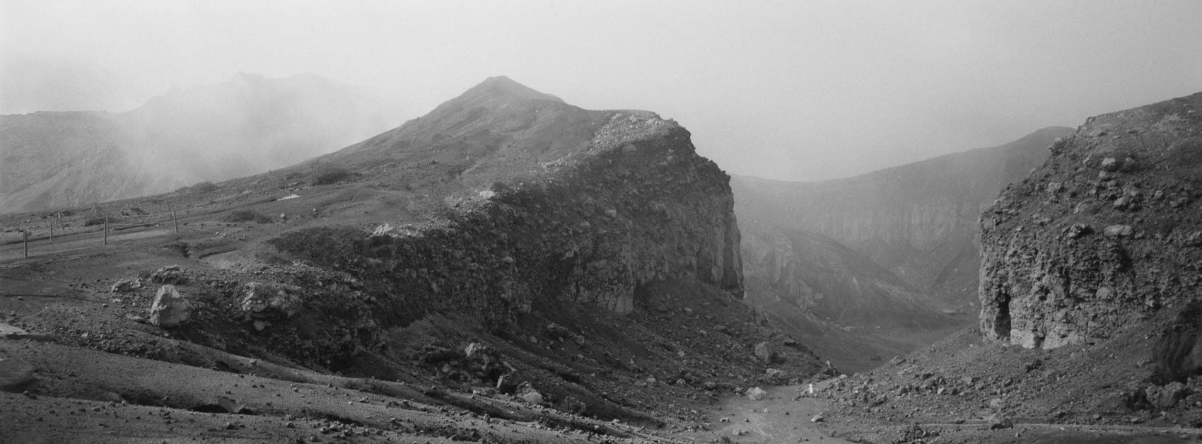 Image alt text: Japan hiking at Mount Aso: an opening in the crater, showing you an entrance to lava in a dramatic black-and-white landscape, with a small fence for scale, captured on Ilford film in Kyushu's otherworldly terrain.
