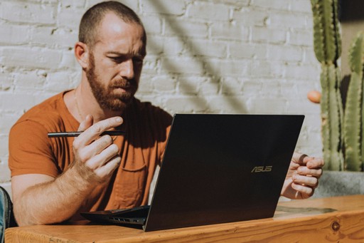 A man with a beard sits at a table using a laptop, holding a cup and looking engaged in conversation.