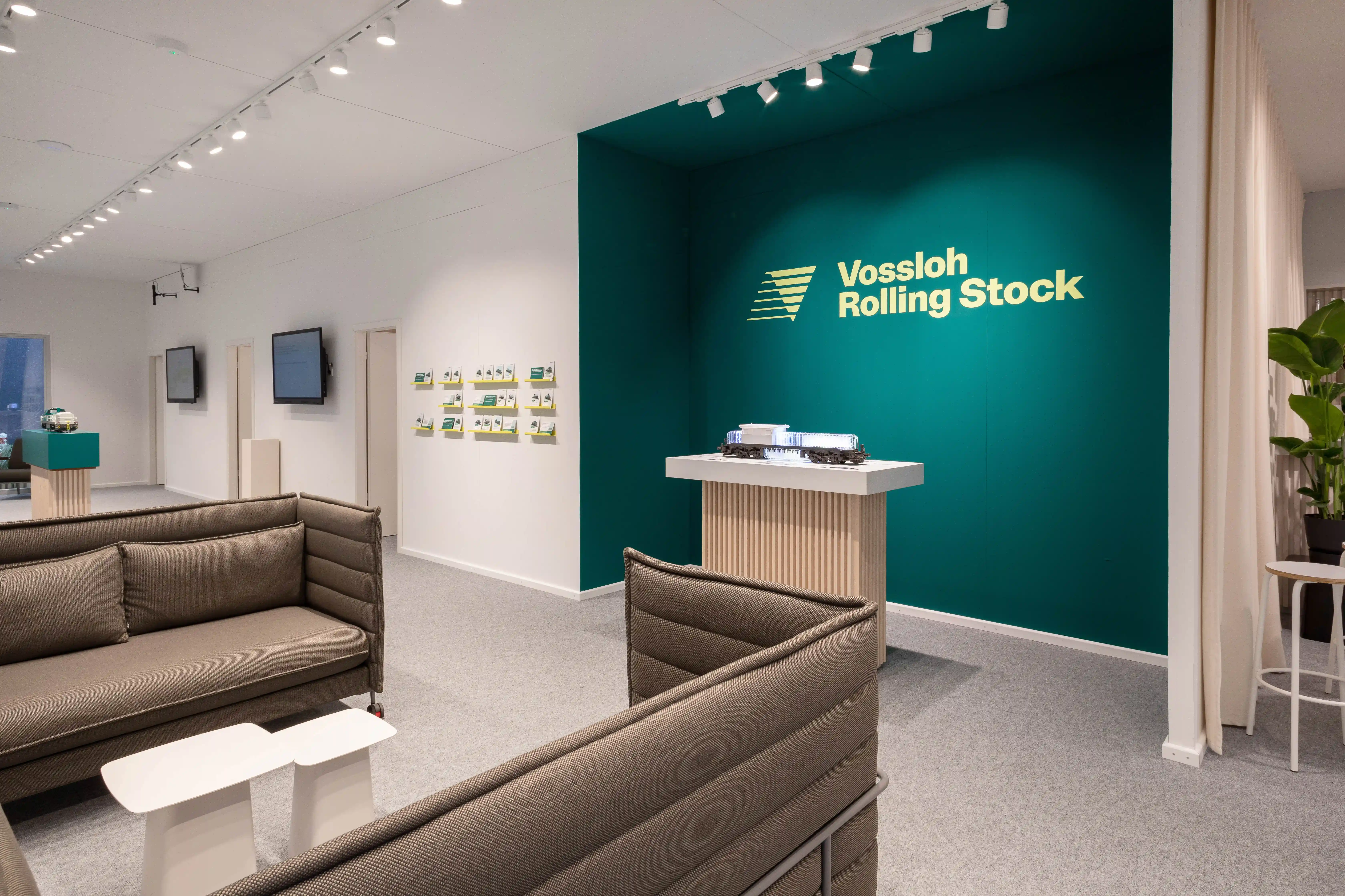 Lounge of the Vossloh house with beige sofas, white side tables and green logo wall with locomotive model.