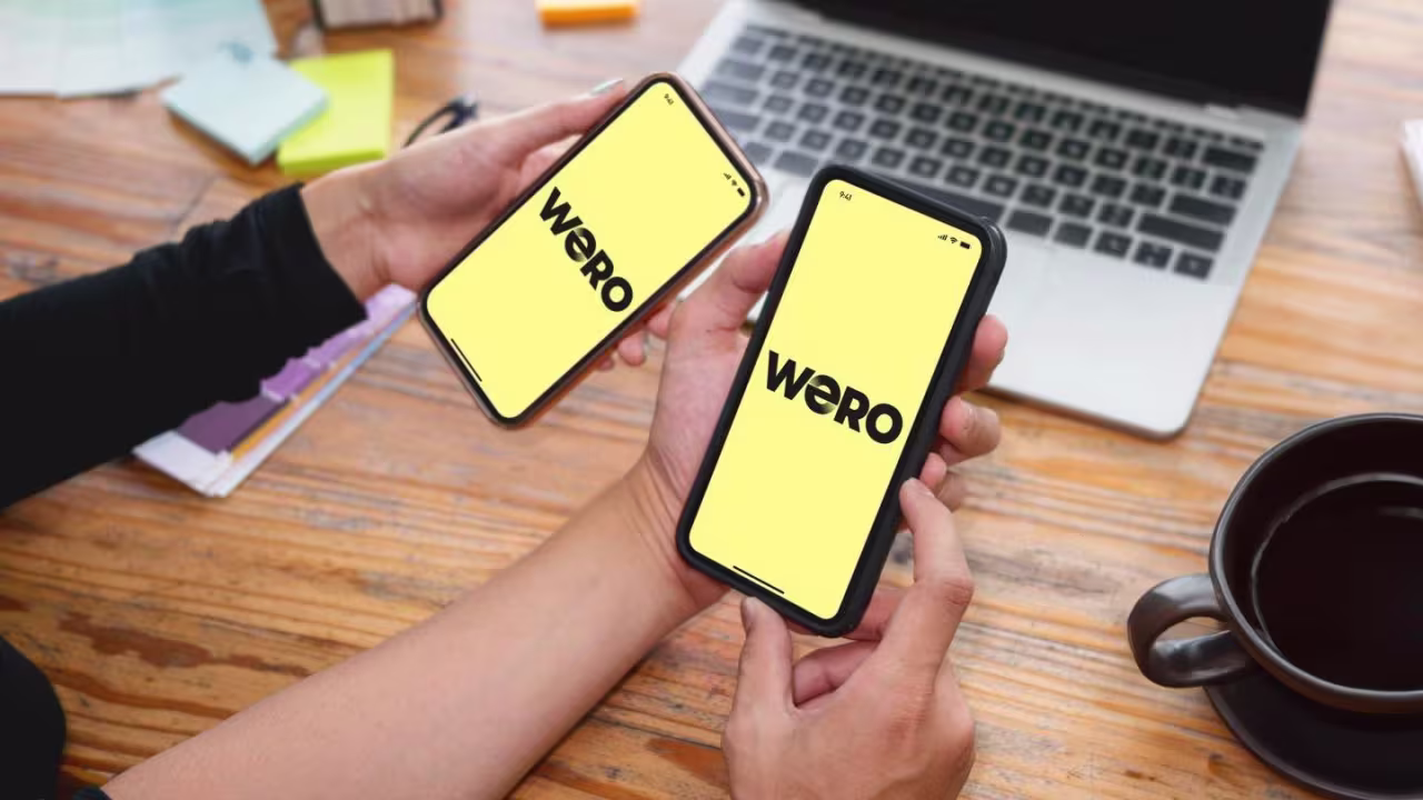Zwei Personen halten Smartphones mit dem WERO-Schriftzug auf dem Bildschirm; im Hintergrund ein Holztisch mit Laptop und Kaffee.