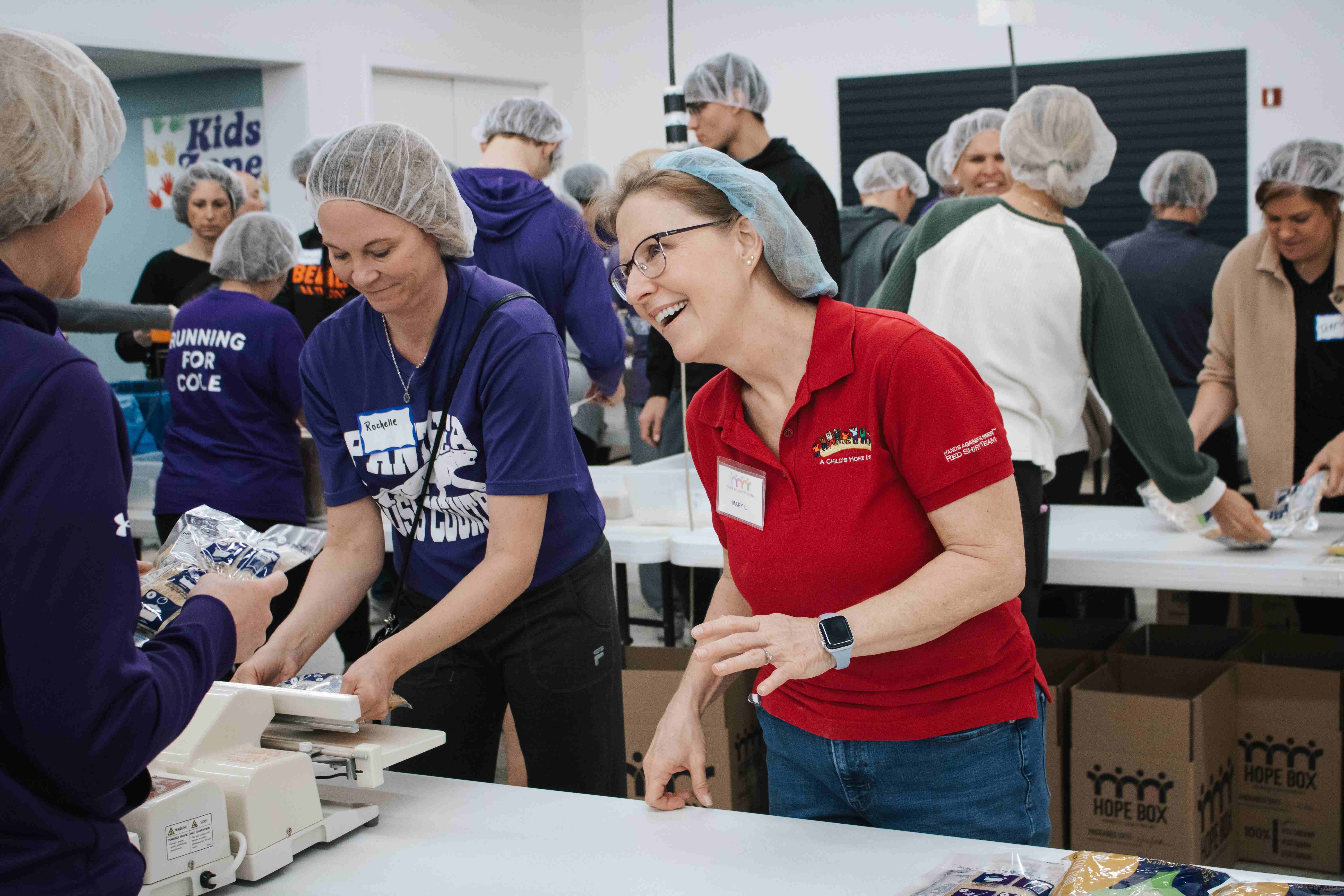 Two women—one in a purple shirt and the other in a red ACHI staff shirt—joyfully packing meals together at a Hands Against Hunger® volunteer event.