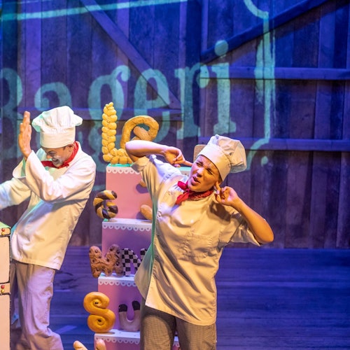 Two chefs in white uniforms and hats perform on stage. One covers his face, and the other covers her ears, with bread props around them.