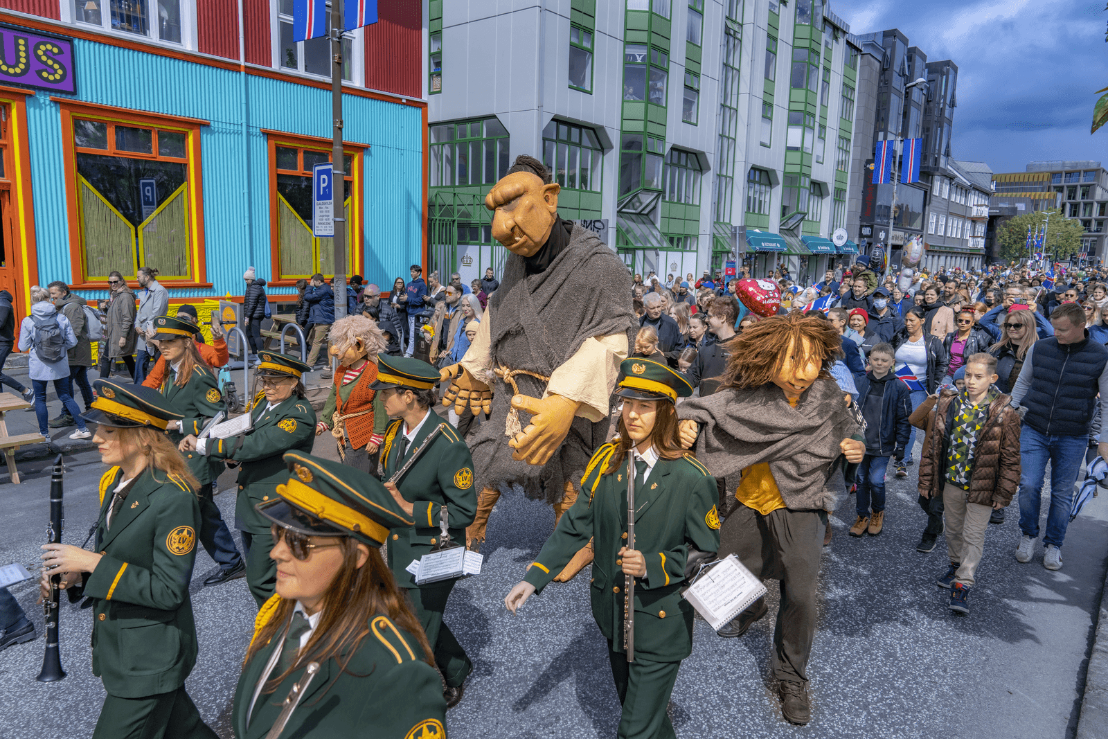 Members of the Lúðrasveit Verkalýðsins marching band, wearing green uniforms with gold trim and peaked caps, play instruments down a crowded Reykjavík street during the June 17 National Day parade. They are accompanied by performers wearing large, expressive troll costumes that interact with the surrounding audience.